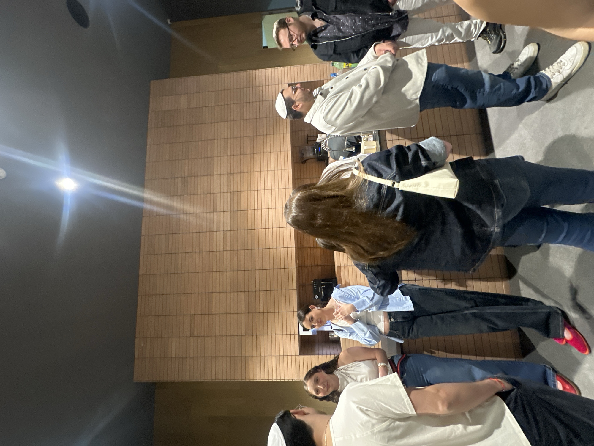 A group of people are standing indoors. They appear to be in a well-lit room with a wall panelled with rectangular wooden tiles. Some people are wearing head coverings. People wear casual clothing, including jeans, jackets, and t-shirts. Their activity is not clear, but they seem to be socializing. No information in the image indicates the location in Huixquilucan de Degollado, Mexico.