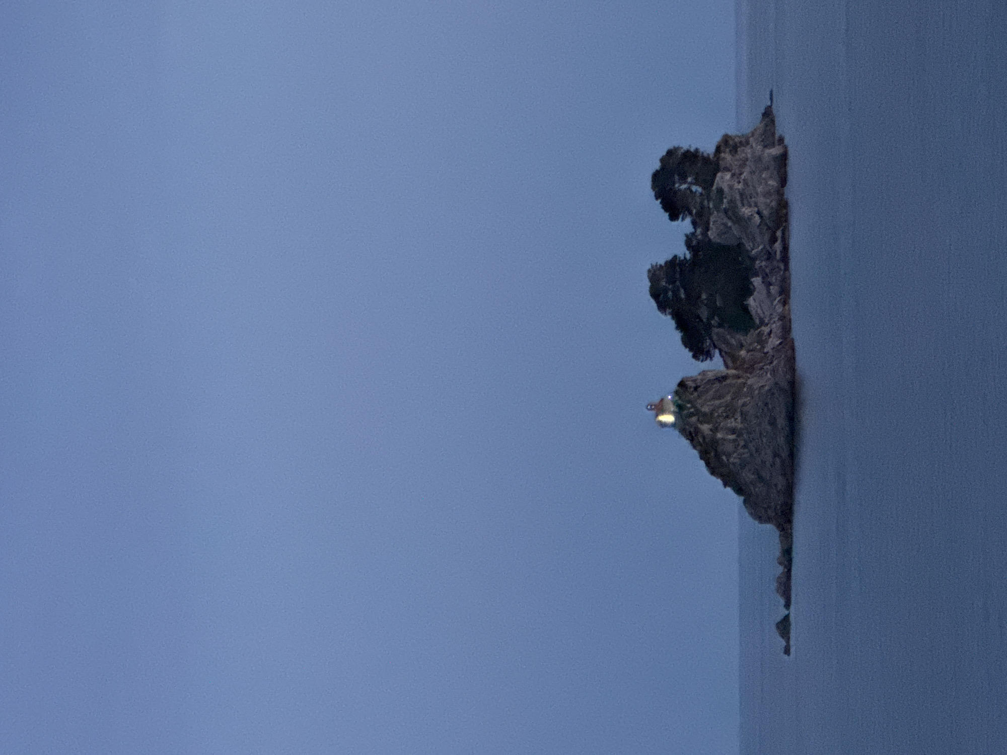 The image presents a rocky islet topped with trees and a small structure that appears to be a lighthouse, situated within a body of water, presumably the Adriatic Sea. The sky above is an overcast pale blue, suggesting either twilight or a heavily clouded day. The composition is such that the islet occupies a small portion of the right side of the frame, with the water extending vertically along the edge and the sky dominating the left side. Gornji Katun, Montenegro is not depicted in this image. The exact location in the image is the Mamula Island.