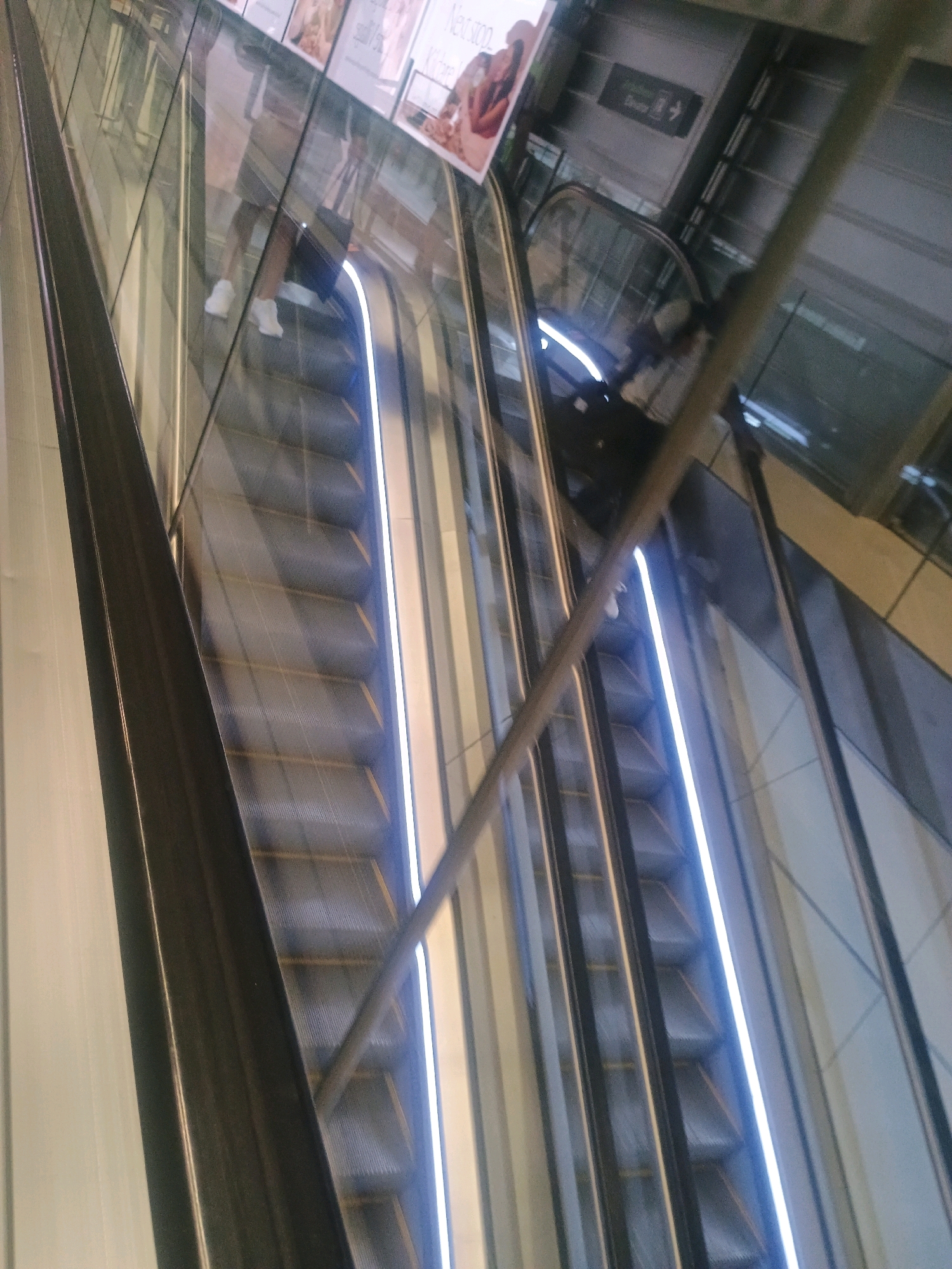 The image captures a close-up, angled view of an escalator in motion, likely within a modern building. The escalator steps are a dark, ribbed material, illuminated by a bright white light strip running along the side of the moving stairs. The handrail, a metallic pole, runs diagonally across the frame. Reflections on the glass panels flanking the escalator show hints of the interior of the building, including what appear to be advertisements or signage. In the foreground, a person's legs are visible, wearing white sneakers and dark shorts, indicating they are ascending the escalator. The overall impression is one of a functional, contemporary urban space.
