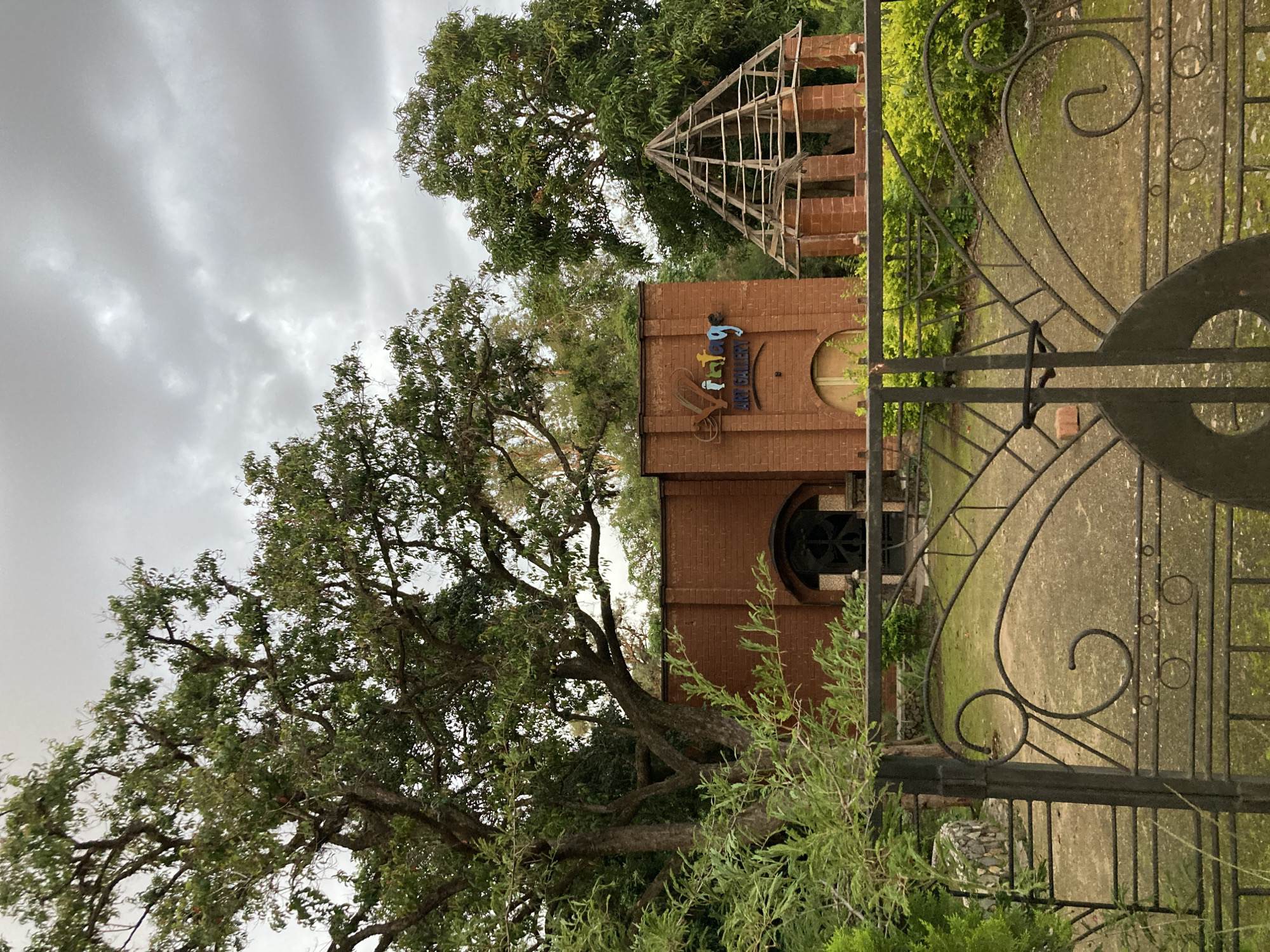 This is a photograph of a brick building with a wooden structure on top, likely an unfinished construction. A black metal gate with decorative scrollwork is in the foreground, partially obscuring the view of the building and its surroundings. Trees with dense green foliage frame the scene, with one large tree in particular dominating the left side of the image. The sky is overcast with heavy, gray clouds, suggesting a cloudy or possibly rainy day. The building has a sign with colorful, stylized lettering that appears to read "ART GALLERY," with "ART GALLERY" in English written below in smaller font. The overall setting seems to be an outdoor space, possibly a garden or park, given the presence of trees and greenery. The time of day is not explicitly clear but the dim light suggests it could be late afternoon or early morning under the cloud cover. There are no people visible in the image, and the mood is somewhat subdued due to the overcast sky and the unfinished construction. The location is identified as Jos, Nigeria.