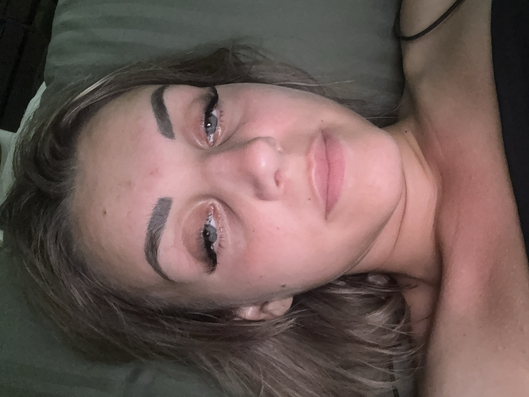 A close-up, top-down selfie of a young woman is shown. She is lying down, facing upwards, with her head tilted slightly. The lighting suggests an indoor setting, possibly a bedroom. Her hair is light brown and falls around her face. She has dramatic false eyelashes and appears to be wearing light makeup. Her skin is fair and appears flushed, particularly on her neck and chest, indicating possible sunburn or redness. She is wearing a black strap top. The background is a dark, textured surface, likely a pillow or bedding, with a hint of a darker fabric behind her head. There are no elements in the image that would allow for localization to Yuzhno-Sakhalinsk, Russia.