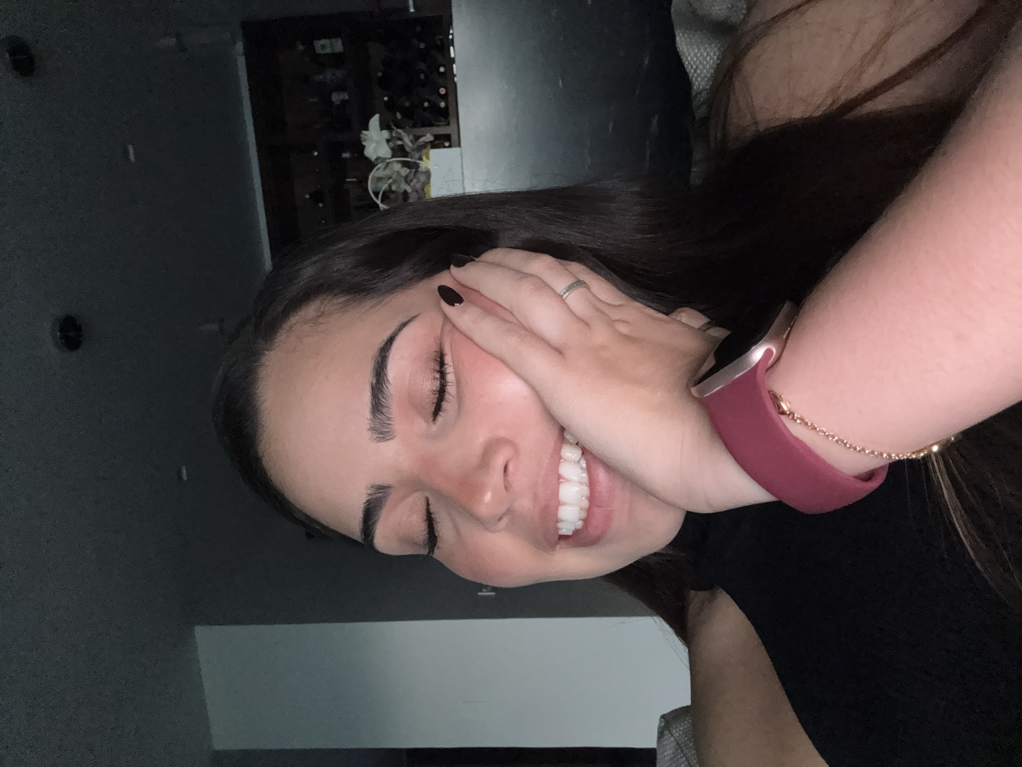 The image depicts a female subject from the chest up, located indoors in San Bernardino Tlaxcalancingo, Mexico. The subject has dark, long hair and fair skin, with a noticeable flush on her cheeks and nose. Her eyes are closed, and her mouth is open in a wide smile, revealing multiple white teeth. Her left hand, featuring black nail polish and a silver-colored ring with a patterned band on the ring finger, rests against her left cheek and jawline. She is wearing a black garment. On her right wrist, a smartwatch with a dark pink band is visible, along with a thin, gold-colored chain bracelet. The background to the subject's left consists of a light-colored wall. To her right, a dark shelving unit or cabinet containing numerous bottles, likely wine, is visible, topped by a white decorative object resembling a floral arrangement. The ceiling is dark and contains recessed light fixtures. Upholstered furniture is partially visible in the lower corners of the frame.