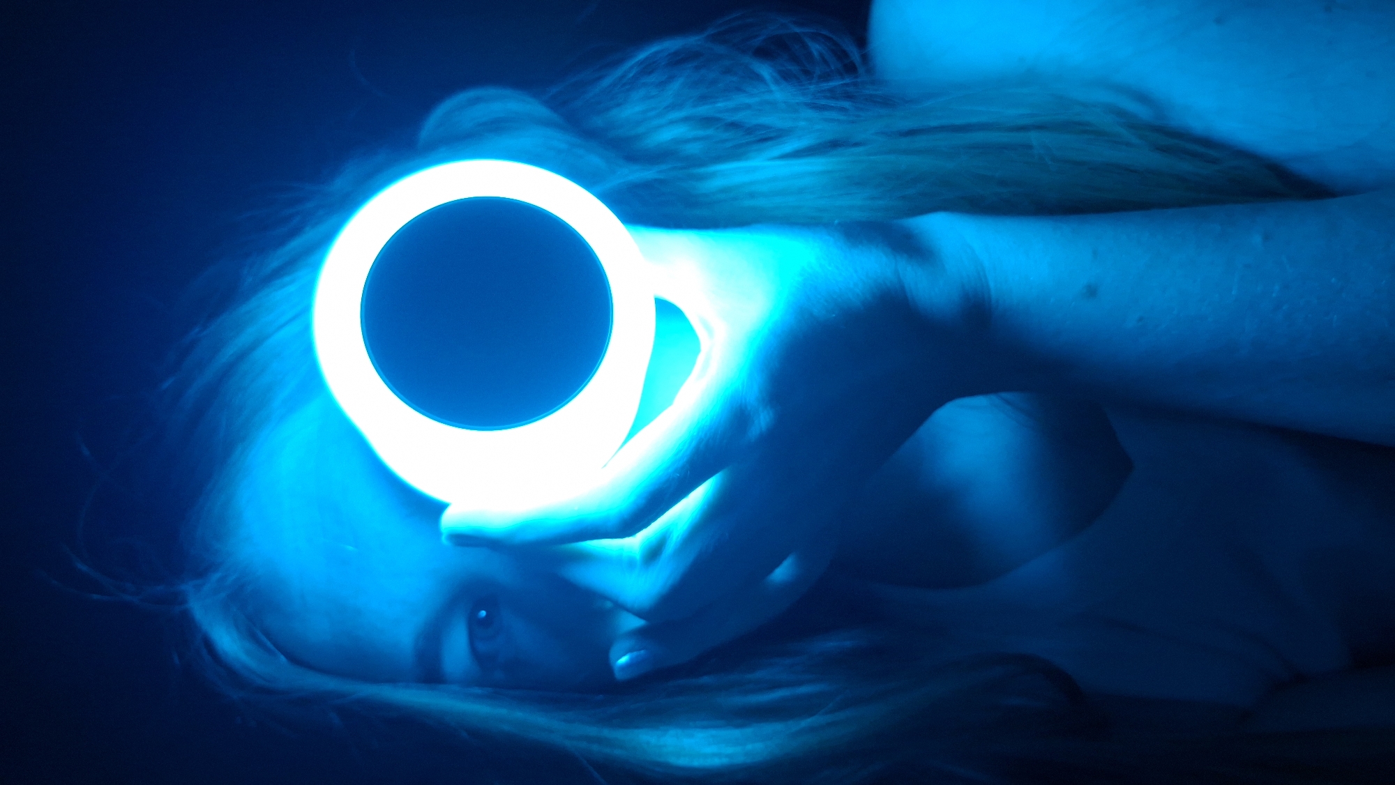 A woman is lying down, illuminated by a bright blue light. She is holding a circular light source directly above her face. Her blonde hair is spread around her head. The background is dark, creating a stark contrast with the bright light. The photograph's subject is the woman, the light she is holding, and the overall blue hue of the scene. No information is discernable to place this in the city of Chaikovsky, Russia.