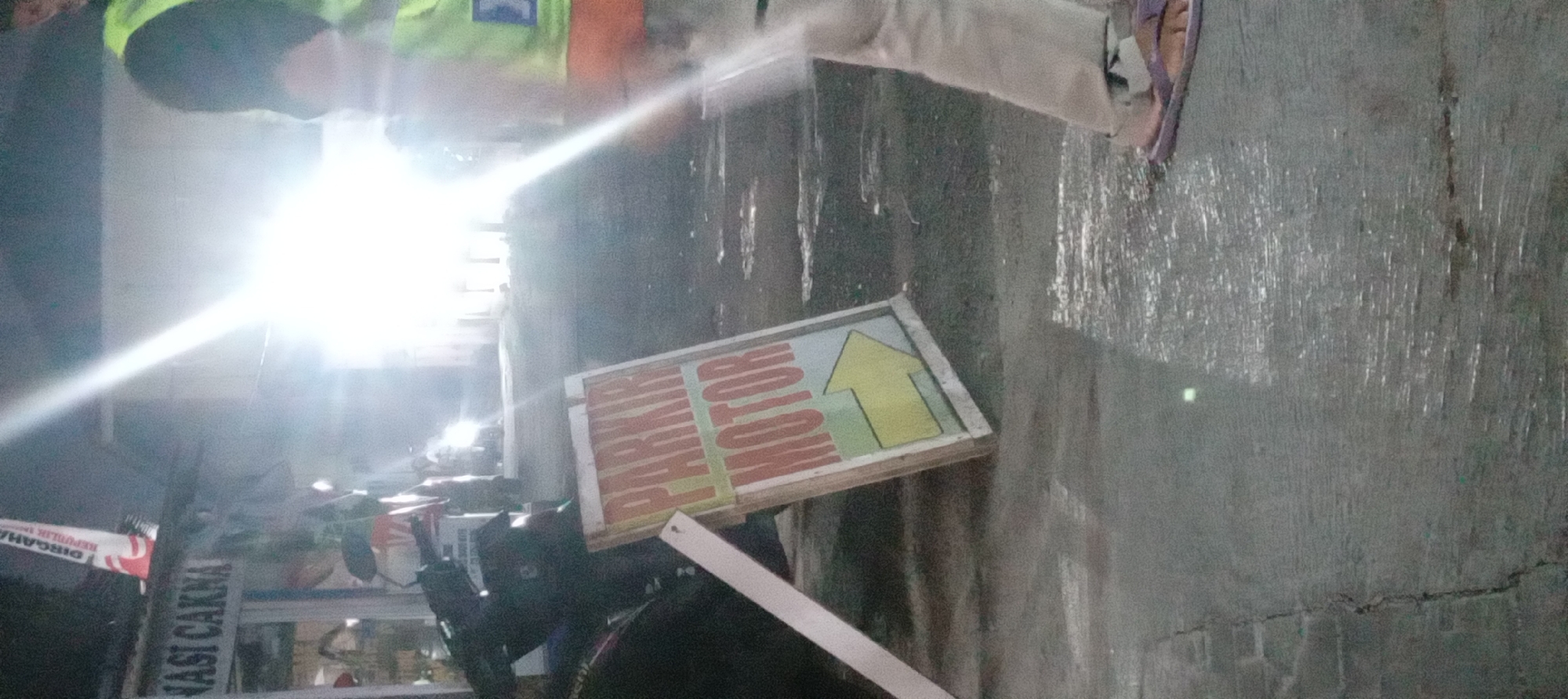 The image shows a street scene at night in what appears to be a commercial area. In the foreground, there is a "PARKING MOTOR" sign with a yellow arrow pointing to the right. A motorcycle is partially visible to the left of the sign. A person wearing a bright yellow vest over a darker shirt and light-colored pants is standing on the right side of the image, facing away from the camera. The person is wearing flip-flops. The ground is wet, suggesting it may have recently rained. In the background, there are lights from buildings and possibly vehicles, creating a bright glare in the center of the image. A banner with Indonesian text, possibly related to a national holiday ("DIRGAHAYU REPUBLIK INDONESIA"), is visible hanging from a building on the left. The overall impression is a busy street scene at night, possibly near a restaurant or shop. While the specific location in Garut, Indonesia, cannot be definitively determined from the image alone, the presence of Indonesian text on the banner and the context of a street scene with a parking sign are consistent with a location in Indonesia.