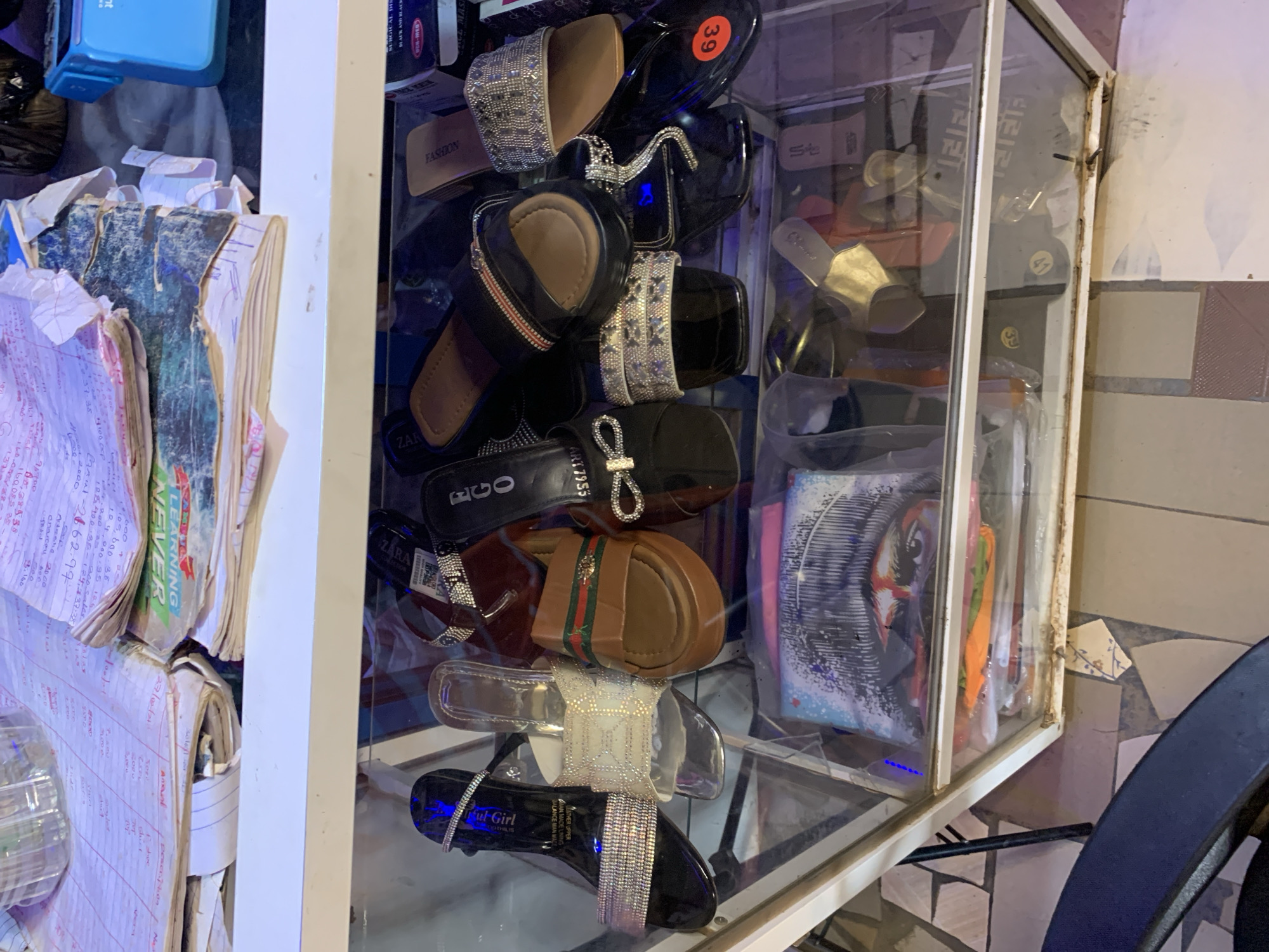 The image shows a glass display case filled with women's sandals. The sandals are various styles and colors, including black, brown, and silver. Some have embellishments like rhinestones. A stack of papers with handwritten notes and a blue stapler are visible next to the display case. The environment appears to be a shop or market stall. The location is Rafin Par, Nigeria.