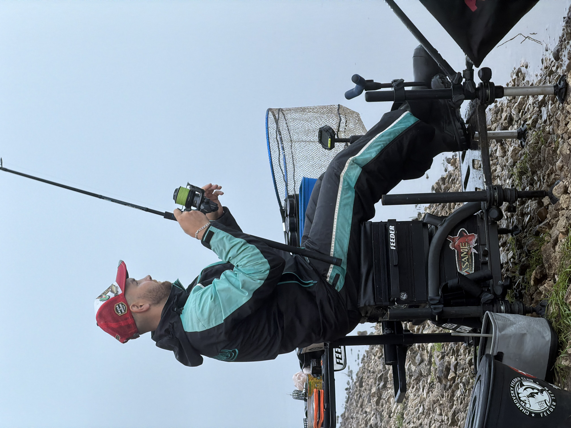 The image, originally rotated 90 degrees, depicts a man engrossed in fishing from a rocky bank beside a body of water under an overcast sky. The angler is seated on a specialized, multi-functional black fishing chair or box, positioned with his back partially towards the viewer. He wears a black and teal jacket, black pants with teal stripes, and a striking red and white baseball cap, which has "CATCH RELEASE" visible on it. His hands skillfully hold a fishing rod, distinguished by a bright green line on its reel, as his gaze appears directed towards the calm water.
The fishing setup is comprehensive, featuring a prominent black feeder box with "FEEDER" text, along with various attachments for rods and equipment. A large landing net is positioned to his side. The ground transitions from small, jagged rocks to the placid, grey water, which extends into a hazy horizon.
Further visible text includes "FEEDER" repeated on different pieces of equipment, "ASAME SUPER CONCEPT JACKIES" on a sticker, and "CHAIKOVSKY FEEDER CLUB TEAM" clearly printed on a black bucket or bag resting on the ground, indicating a specific fishing club from the Chaikovsky, Russia region. The muted light from the cloudy sky contributes to a focused atmosphere, highlighting the specialized gear and dedication involved in the sport of feeder fishing.
