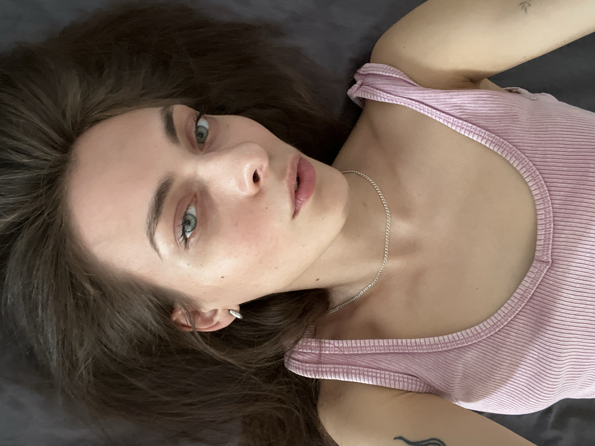 A young woman with light skin and brown hair is lying on a dark gray surface, possibly a bed or floor. She is wearing a pink tank top with a white striped pattern and a silver chain necklace. A small tattoo is visible on her left arm near the shoulder. Her eyes are light-colored, and she is looking directly at the camera with a neutral expression. An earring is visible in her left ear. The location is not discernible from the image.