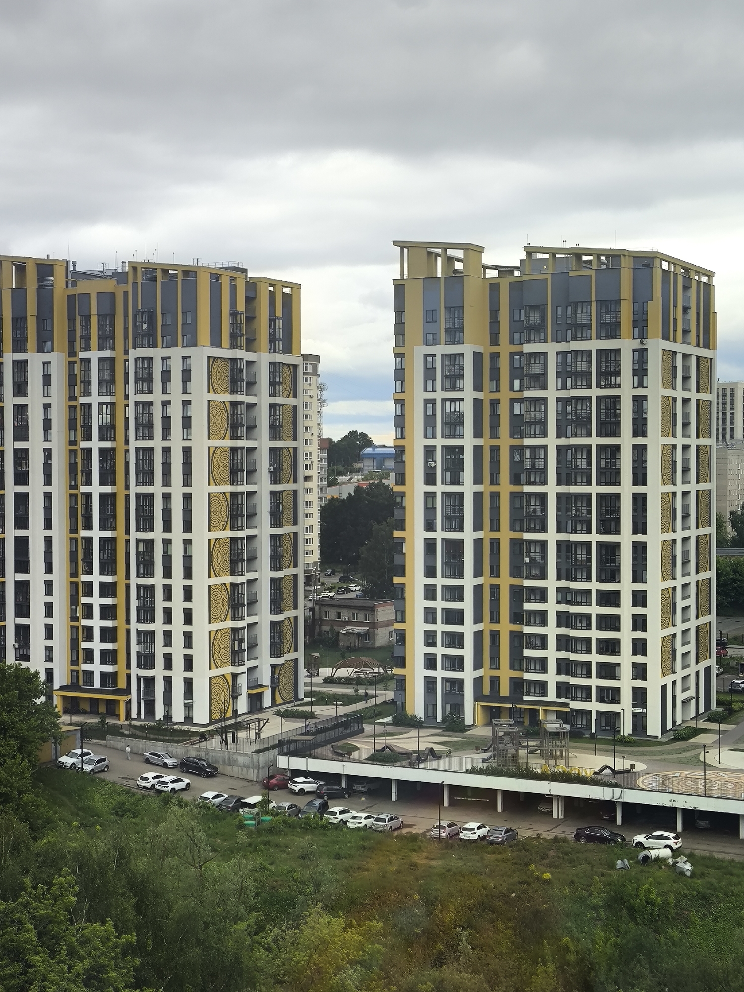 The image displays an elevated view of a modern urban residential complex in Unknown, Unknown, under an overcast sky. Two prominent, multi-story residential buildings, identical in design, stand centrally. These towers feature contemporary architecture with facades composed of white, dark gray, and yellow/gold panels, arranged in vertical and horizontal blocks. Distinctive gold circular patterns decorate several vertical white sections of the buildings. The rooftops are flat with decorative yellow/gold and dark gray architectural elements and small antennas.
At the base of the buildings, an elevated concrete platform hosts a children's playground with yellow and dark gray equipment and multiple lamp posts. Below and around this structure, various paved driveways and open-air parking areas are visible. Numerous passenger cars, including white, silver, black, and red vehicles, are parked along the roads and in designated spaces, some partially beneath the elevated platform. A single individual in light-colored clothing walks on a path near the base of the left building.
The foreground is occupied by a dense area of lush green trees and bushes, suggesting a natural slope or parkland. Additional trees and glimpses of other, more distant buildings are visible in the background and between the two main towers. The sky is uniformly gray and cloudy, providing diffused lighting to the entire scene.