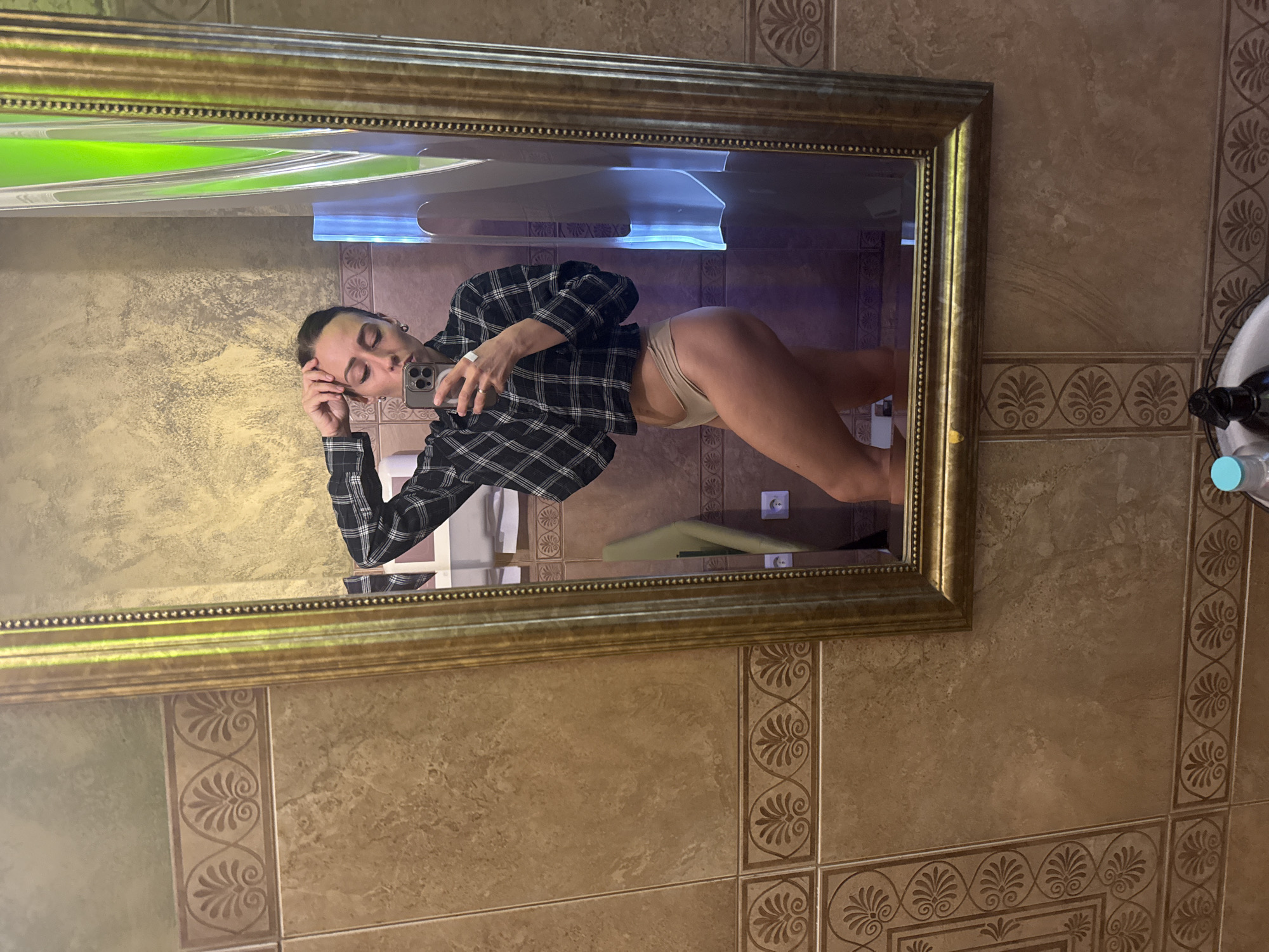 The image, rotated 90 degrees counter-clockwise, displays a mirror selfie taken by a person in an indoor setting, likely in Saint Petersburg, Russia. The central subject is a female-presenting individual, visible from the waist up in a large, ornate gold-framed mirror. She is wearing a black and white plaid shirt, unbuttoned, revealing light-colored (possibly beige) high-cut underwear bottoms. Her hair is pulled back, and her eyes appear closed or looking down as she holds a silver smartphone (likely an iPhone with multiple cameras) in her right hand to capture the reflection. Her left hand rests casually on her head.
The background of the reflection reveals light-brown or tan tiled walls with decorative borders featuring repeated patterns, possibly floral or Greek key-like motifs. A white electrical outlet is visible on the wall to the right of the person's reflection. Bright reflections of green and blue light sources are present in the upper portion of the mirror. On the far right edge of the image, a partial view of a bottle or dispenser with a light blue cap is visible. The scene suggests a private space such as a bathroom or dressing room. No visible text is discernible in the image. The time of day is not indicated.