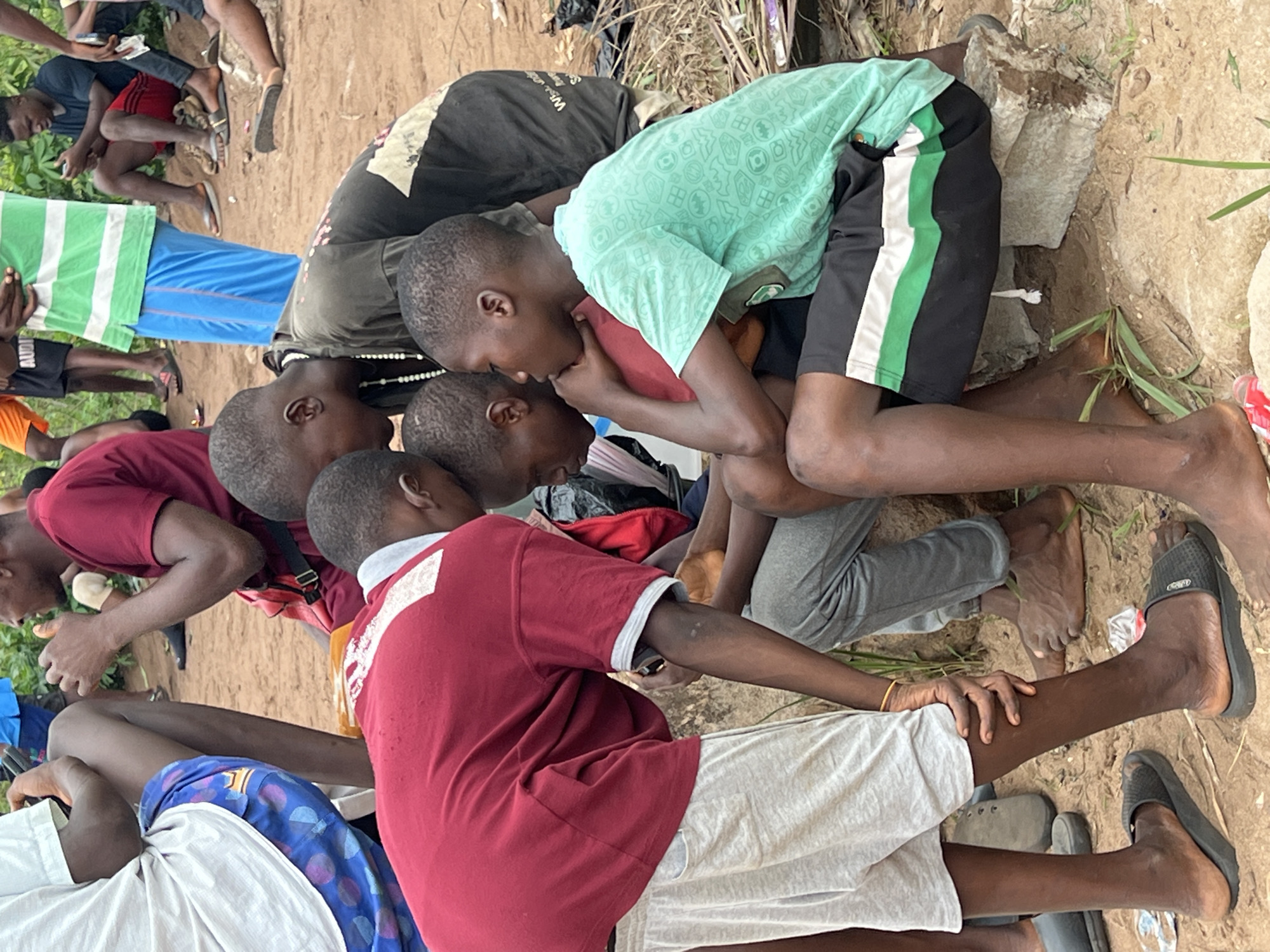 In Nkpor, Nigeria, a group of people huddle together. The individuals are predominantly children and young adults with dark skin, dressed in casual clothing. One person wears a green shirt with black shorts, while another is in a red shirt and white shorts. The group is gathered on a dirt surface with scattered debris, including what appears to be garbage and rocks. Some people are barefoot, while others wear sandals. The overall environment suggests a low-income or informal setting.