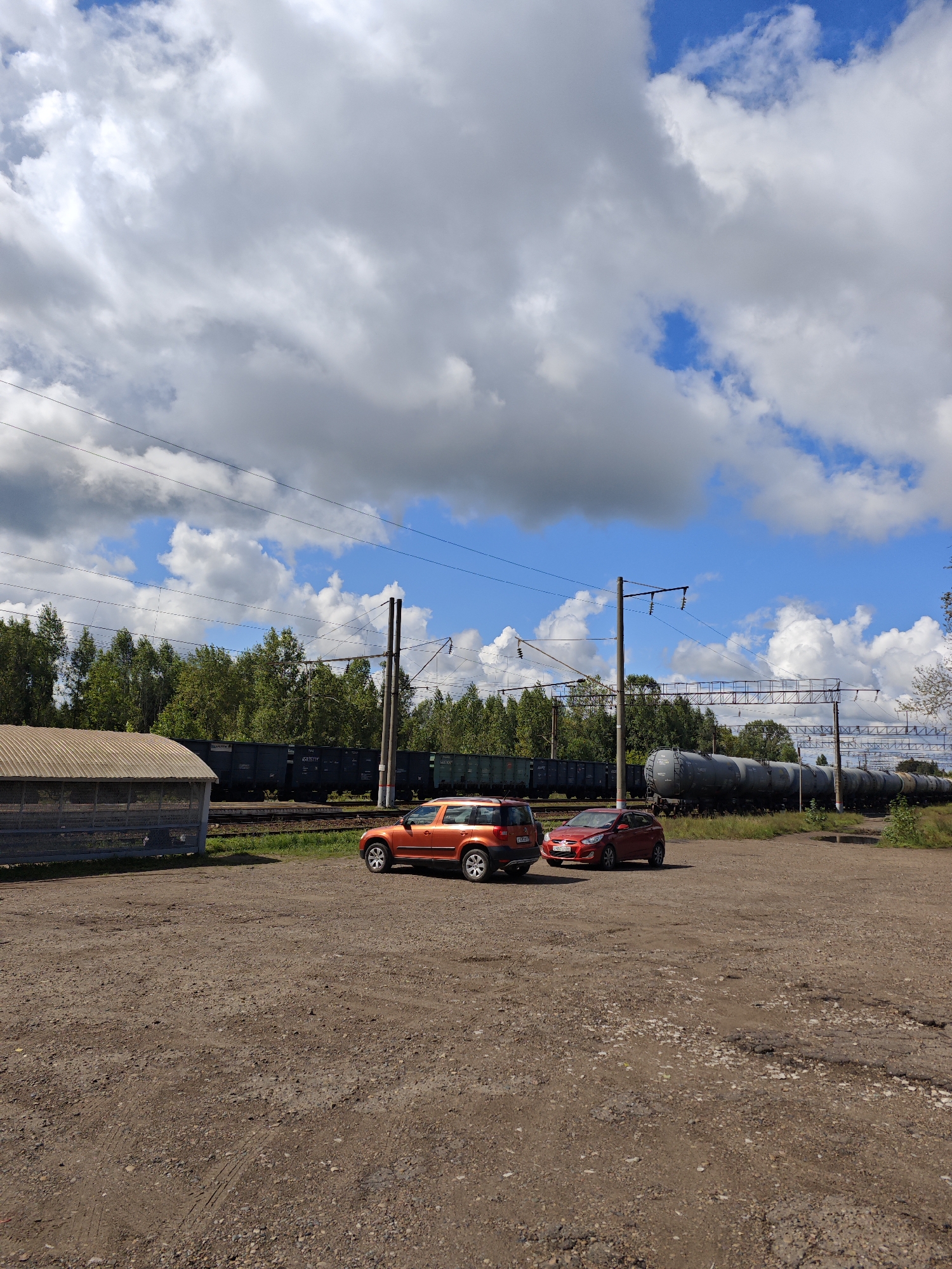 In Kambarka, Russia, two cars, an orange SUV and a red hatchback, are parked on a dirt lot with a large building to the left. In the background, a long train of dark freight cars and tank cars is visible, traveling on railway tracks. The train is flanked by a dense line of green trees. Overhead, a bright blue sky is filled with large, white, fluffy clouds. The overall scene suggests a sunny day at a transportation hub.