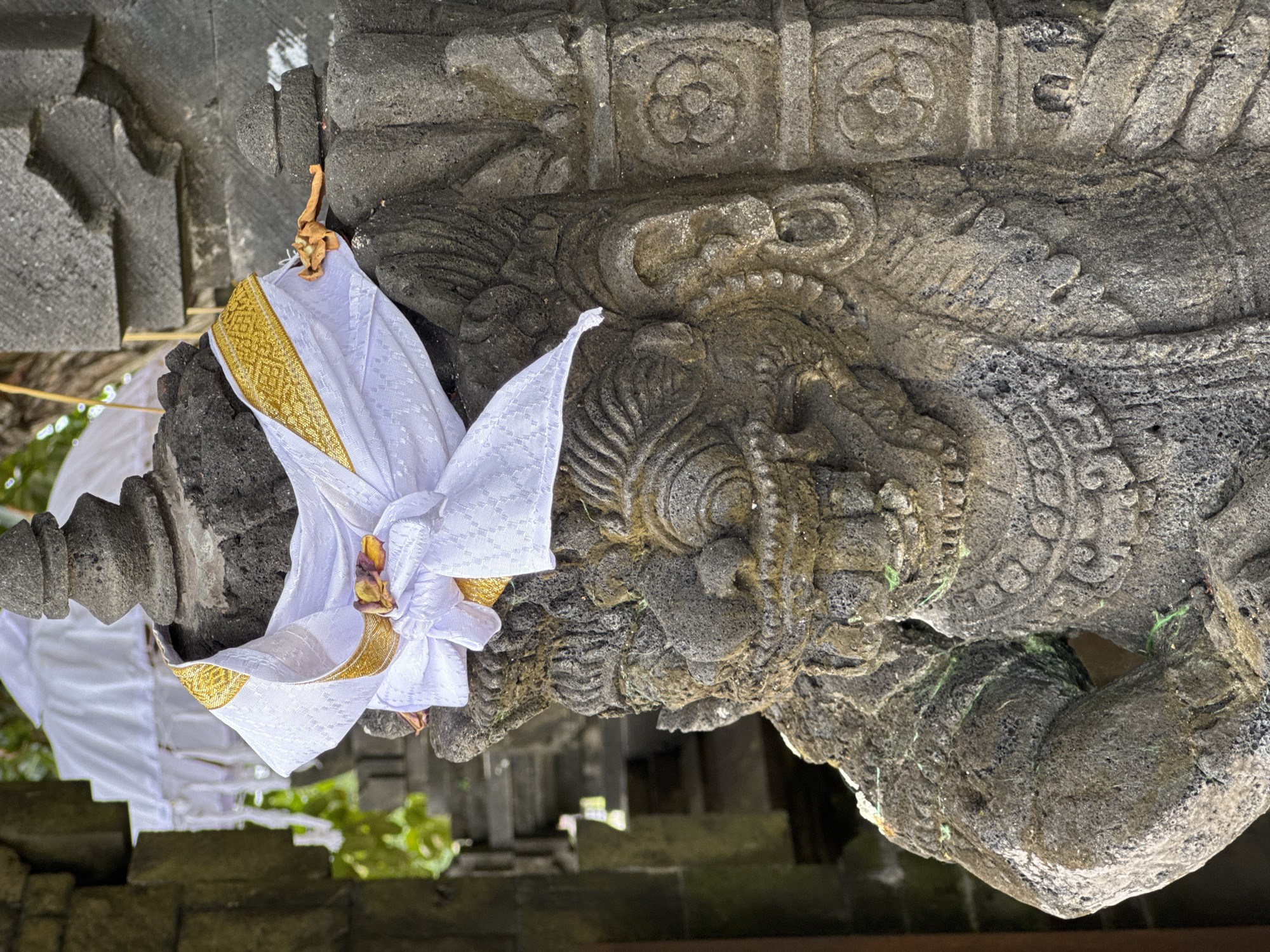The image captures a close-up view of a weathered stone statue, likely a guardian figure, located in Kuta Selatan, Indonesia. The statue is adorned with a white fabric sash featuring gold trim, tied around its upper body. The fabric is intricately folded and knotted, with a small, dried flower tucked into the knot. The stone carving depicts a fearsome face with prominent teeth, wide eyes, and elaborate headdress. The texture of the stone is rough and aged, with some patches of green moss growing on its surface. In the background, glimpses of stone architecture and greenery suggest a temple or religious site. The lighting indicates it is daytime, with shadows cast on the statue.