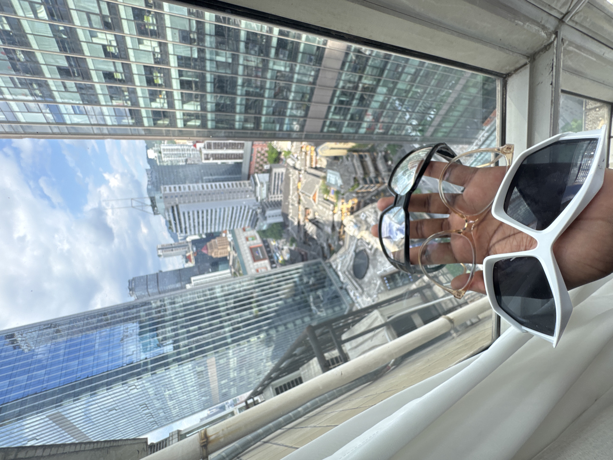 A person's hand, holding three pairs of eyewear, is positioned in the foreground of the image, obscuring the lower portion of the scene. Two pairs of spectacles, one with black frames and clear lenses, and another with gold-toned frames and clear lenses, are held open. A third pair of oversized white sunglasses with dark lenses rests on the palm. Behind the hand, a panoramic view of a dense urban cityscape unfolds. Towering glass skyscrapers dominate the background, their facades reflecting the sky and surrounding buildings. Below the skyscrapers, older, more traditional architecture with gray tiled roofs is visible, suggesting a mix of modern and historical urban development. The scene appears to be captured from an elevated vantage point, possibly a high-rise building, with a window frame visible in the lower right corner. The overall impression is one of a bustling metropolis with a distinct architectural contrast. The location is Yuzhong District, China.