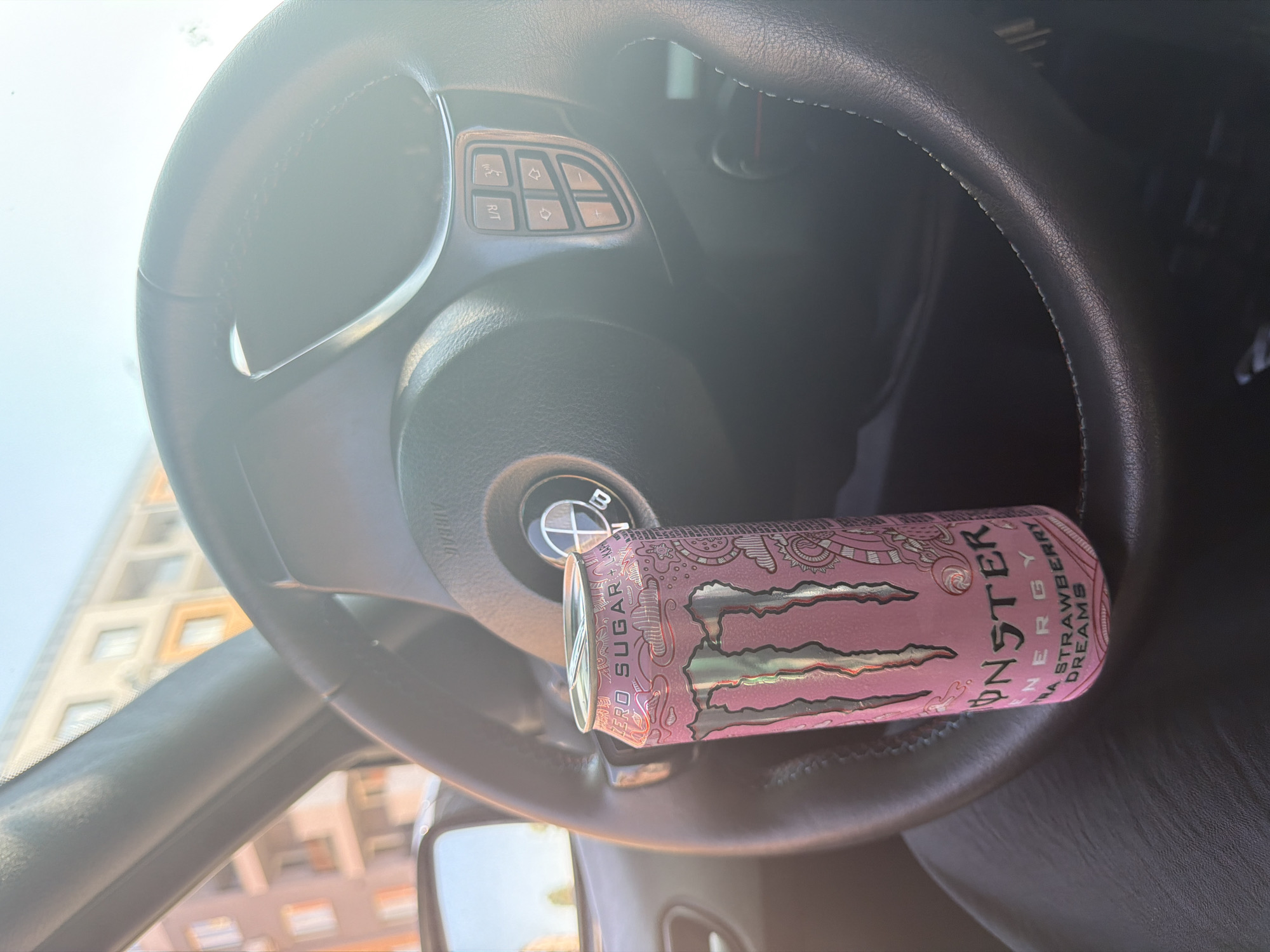 An eye-level shot captures a pink Monster Energy drink can resting horizontally on a black leather steering wheel inside a car, likely in Pazardzhik, Bulgaria. The can is prominently labeled "MONSTER ENERGY ULTRA STRAWBERRY DREAMS" and "ZERO SUGAR". The steering wheel features a partial BMW logo on its center airbag cover and integrated controls on the right spoke, including phone call icons, volume up/down, and an "R/T" button.
The scene is brightly lit by strong sunlight, causing significant glare on the dashboard and windshield, suggesting a clear, sunny daytime. Through the windshield, part of a multi-story building with cream-colored and orange-framed windows is visible against a bright, clear sky. The overall impression is a casual moment inside a parked vehicle.