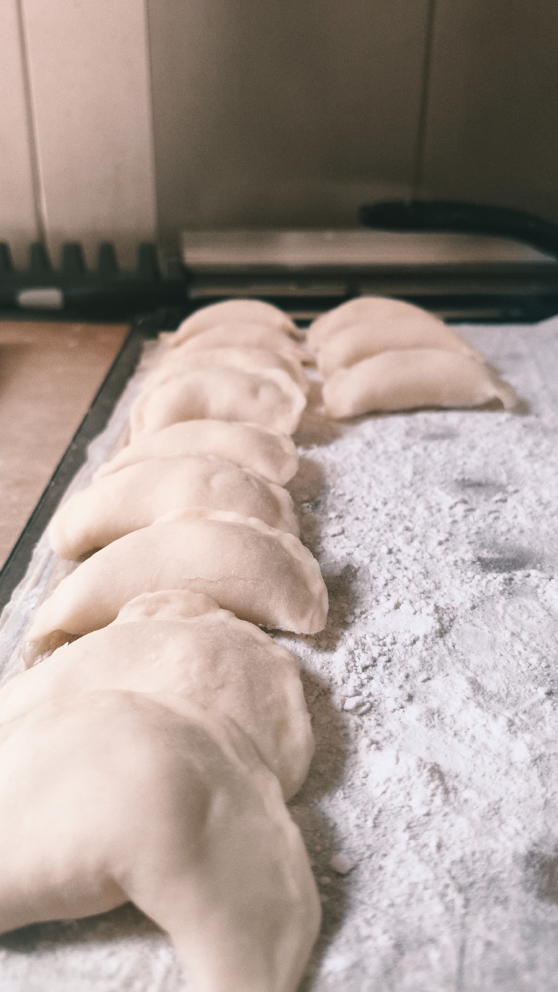 The image shows a close-up view of a row of uncooked pierogi or dumplings arranged on a flat surface heavily dusted with white flour. The pierogi are light in color, likely made from dough, and have a crescent shape with crimped edges. The surface appears to be a baking sheet or countertop. The background features a light-colored tile wall and a dark appliance. The image was likely taken in a kitchen. The location in Nowy Tomyśl, Poland is not discernible from the visual information alone.