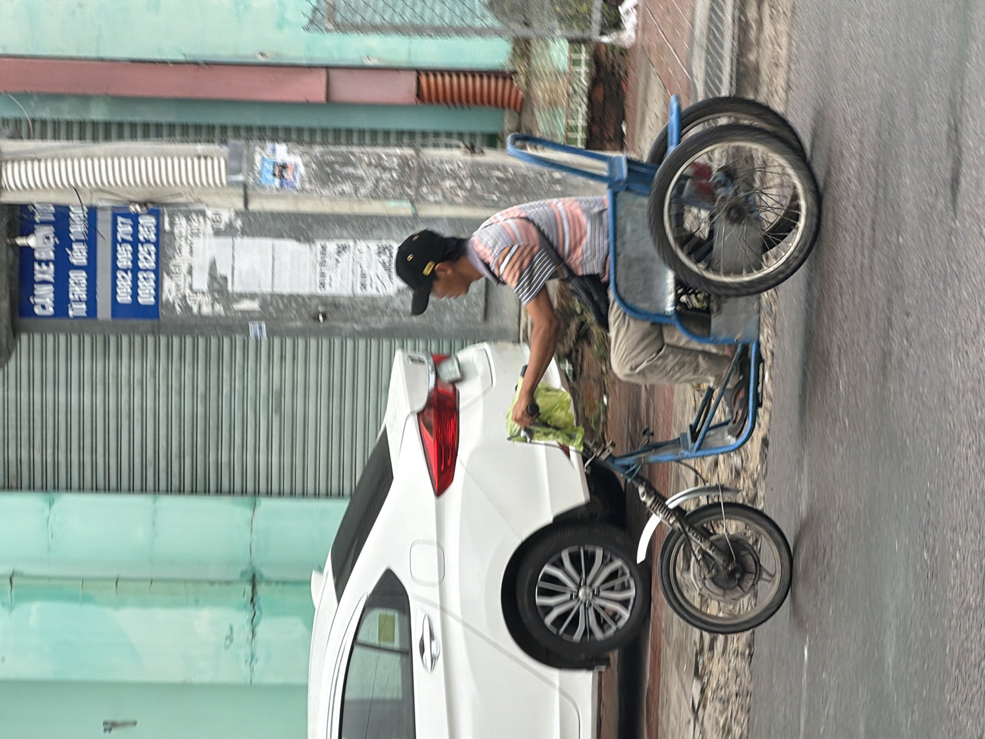 An individual wearing a black baseball cap, a striped short-sleeved shirt, and light-colored pants is riding a three-wheeled vehicle with a blue frame. The person is holding a green cloth in their right hand. The vehicle has two wheels in the rear and one in the front. To the left of the three-wheeled vehicle is a white sedan. The scene appears to be on a paved street, with a building featuring a turquoise-colored wall and a corrugated metal gate in the background. A utility pole with blue and white signage, indicating "CỬA HÀNG XE ĐIỆN" (Electric Bike Store) and phone numbers, is also visible. The image was captured in Nha Trang, Vietnam, indicated by the signage and the context of the scene. The lighting suggests an overcast or cloudy day.