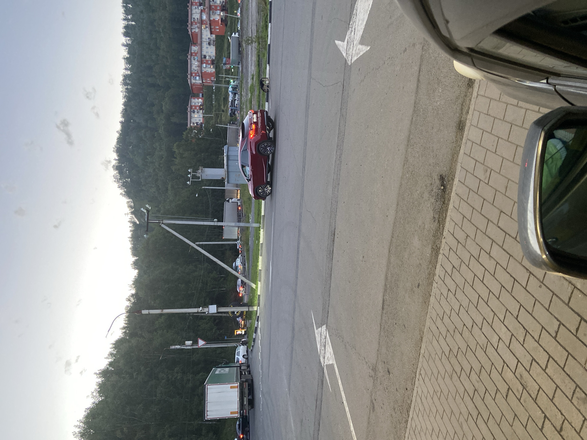 In the city of Slyudyanka, Russia, a suburban street scene unfolds under an overcast sky. A red Lexus RX SUV is stopped on the right side of the road, facing left, with its brake lights illuminated. A dog is sniffing the ground near the curb beside the SUV. Across the road, a large white truck with a green stripe on its trailer is also stopped, facing right, with its hazard lights on. Several other vehicles are visible in the background, indicating moderate traffic. Residential buildings with red roofs are situated on a tree-covered hillside in the distance. The foreground shows a brick-paved sidewalk. The overall impression is that of a roadside scene in a town or city, possibly near a junction or a service area.