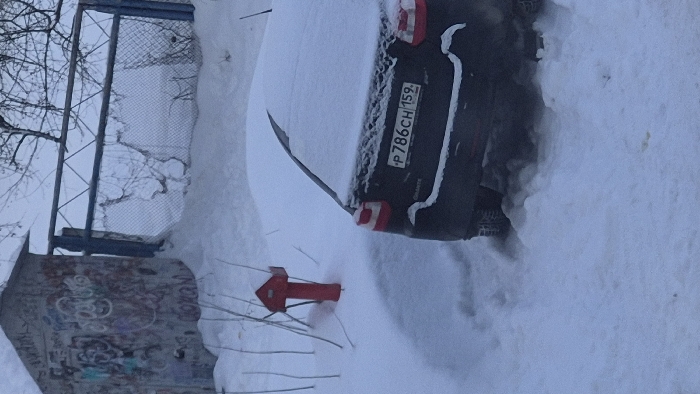 A car, partially buried in snow, is parked next to a red fire hydrant, also partially covered in snow. The car's license plate is visible: "P786CH 159". A chain-link fence and a small building covered in graffiti stand nearby. The entire scene is blanketed in snow, suggesting a recent heavy snowfall. Bare tree branches are visible in the background.