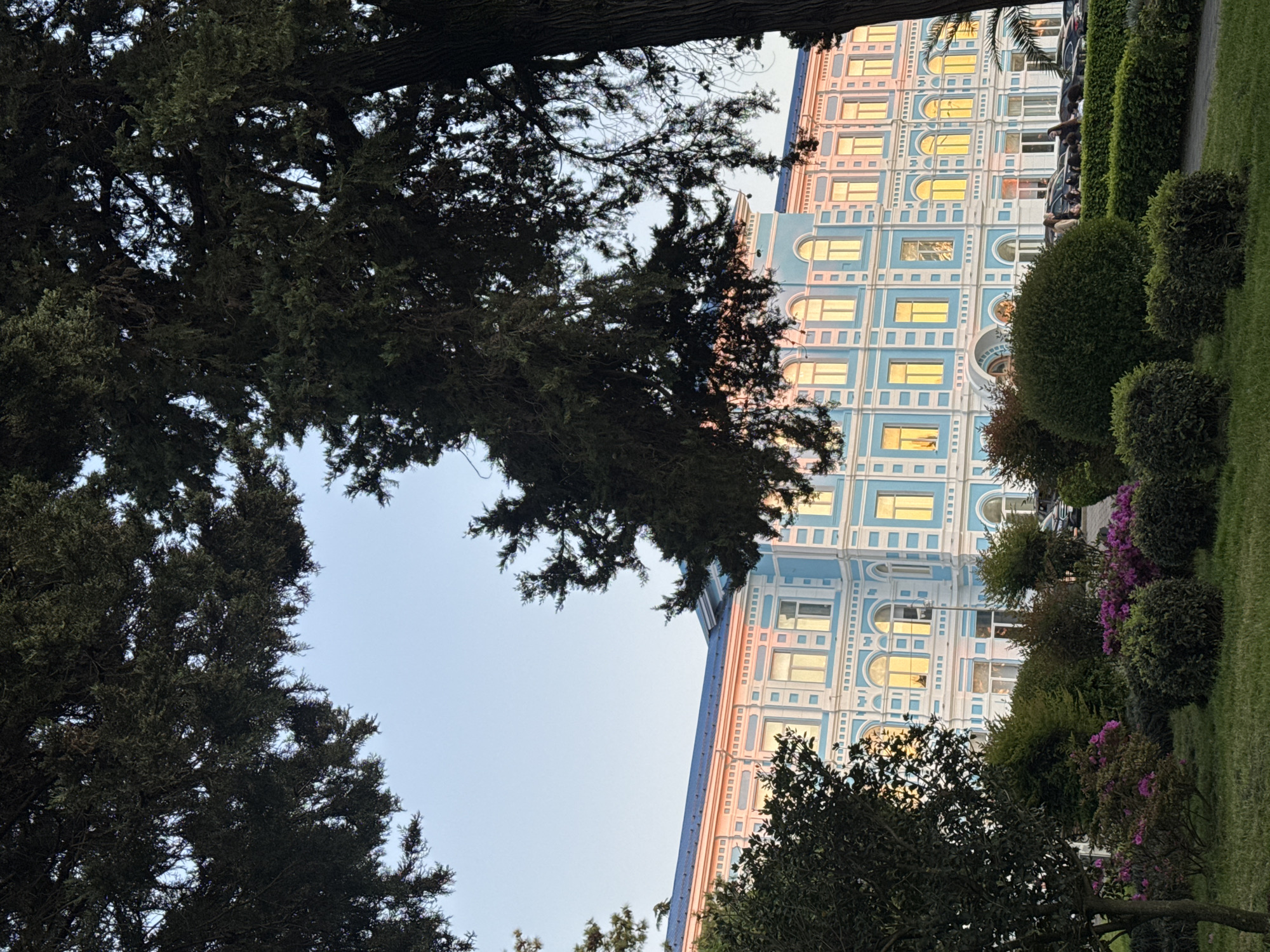 Captured from a low angle looking upward, the image presents a building with a facade painted in blue and white, interspersed with numerous windows reflecting a golden light, indicative of either sunrise or sunset. Flanking the building's right side, a meticulously maintained green hedge stretches, interspersed with blooming pink flowers, suggesting a garden or park-like setting. To the left, a dense canopy of trees frames the view, obscuring the sky, yet contributing to the naturalistic feel of the urban landscape. The photograph exhibits a balance between the architectural and natural elements, implying an urban setting integrated with green spaces.