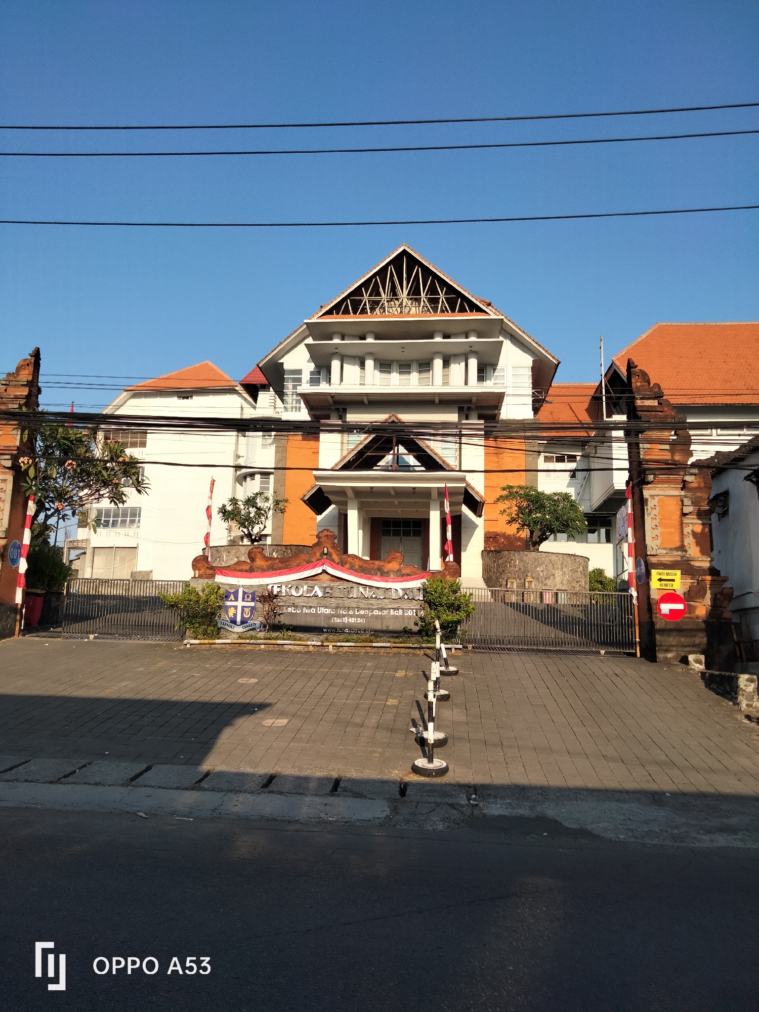 The image shows the entrance to a school building in Denpasar, Indonesia. The school is named "SEKOLAH TUNAS DAUD" and is located at "Kebo Iwa Utara No.8 Denpasar". The building is multi-storied with white and orange walls and a red tiled roof. The entrance features traditional Balinese gates made of brick. A metal gate is visible in front of the building. Several trees and plants are present in the foreground and on the grounds. The sky is clear and blue, suggesting a sunny day. In the bottom left corner of the image, a logo with "OPPO A53" is visible, indicating it was taken with an Oppo A53 phone.