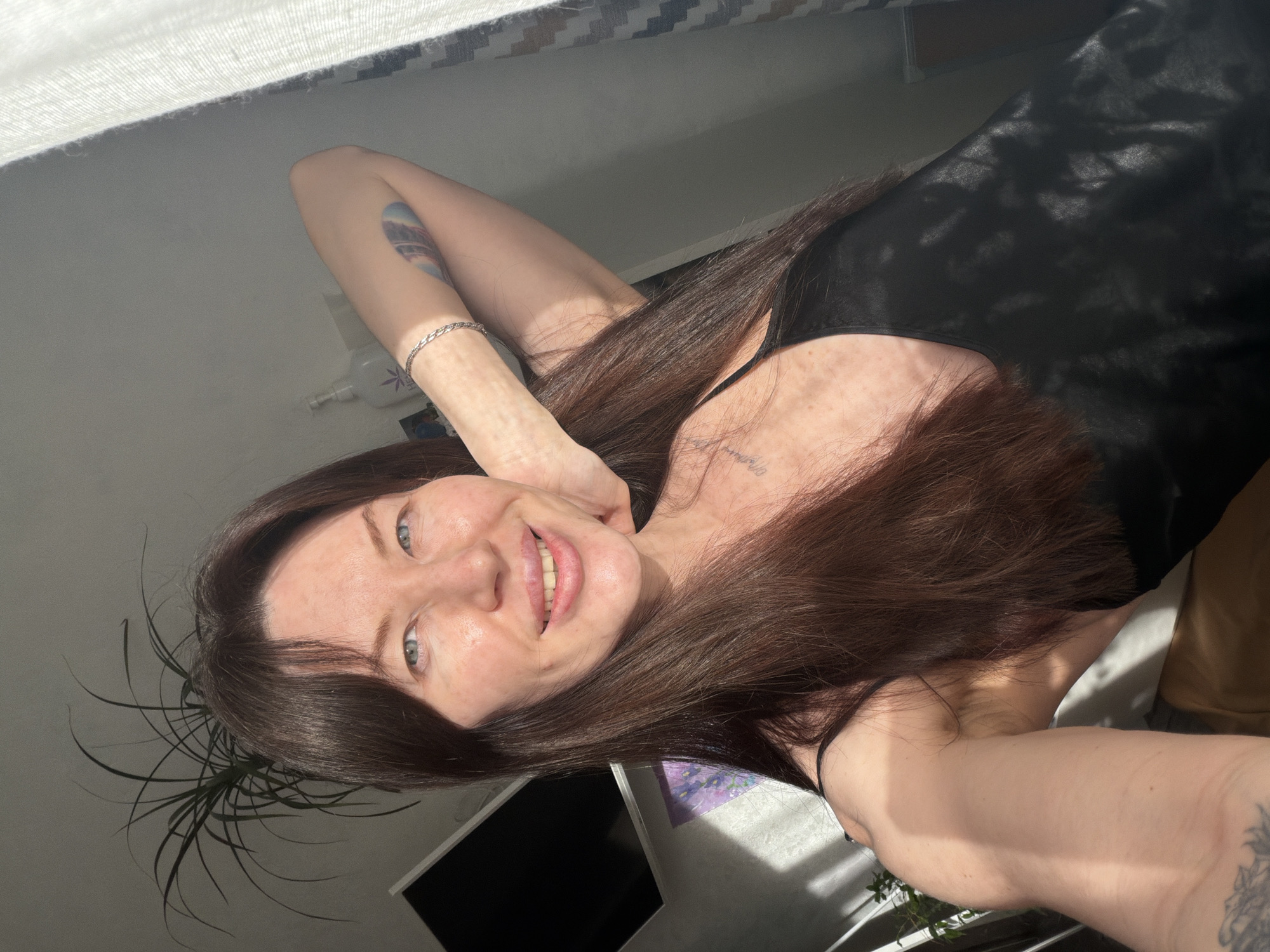 A woman is lying down taking a selfie in what appears to be an indoor setting with a window, a television and a plant visible in the background. The woman has brown hair, blue eyes, and is wearing a black top. Her right arm is bent behind her head. She has a tattoo on her left forearm, a silver bracelet on her left wrist, and another tattoo on her chest. The location could be Perm, Russia, based on the prompt, but is not discernible from the image itself.