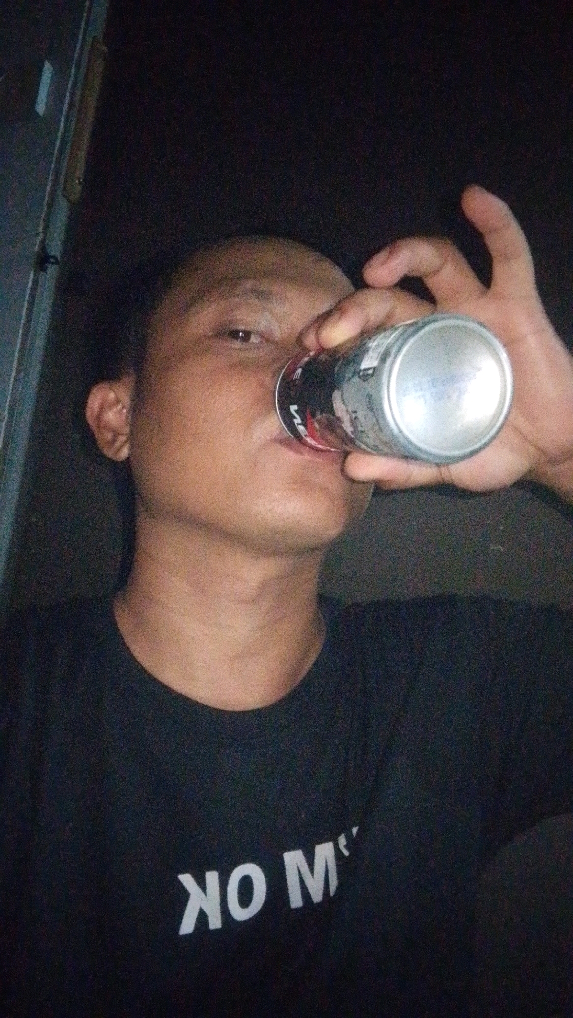 In the dimly lit outdoor setting of Cintaratu, Indonesia, a close-up shot captures a person from the chest up, holding a silver can and drinking from it. The person, facing the camera, has their right hand on the can. They are wearing a black t-shirt with white text that appears to read "NO M". The background is dark and indistinct, suggesting nighttime or a poorly lit area, with a vertical metallic element visible on the left side of the frame.
