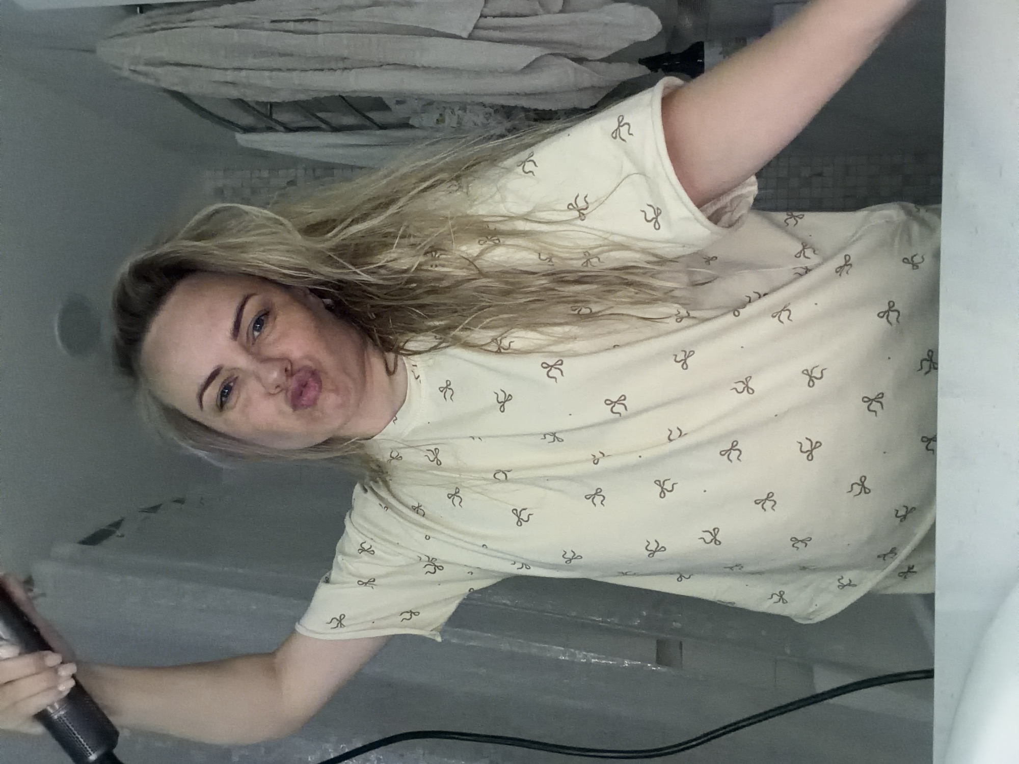 Here's a detailed description of the image:
**People:** The image features a woman with blonde hair and blue eyes. She is holding a hair dryer and appears to be taking a selfie. Her lips are pursed.
**Subjects:** The primary subject is the woman herself. The hair dryer is also a significant subject.
**Objects:** Objects present in the image include the hair dryer (held by the woman), a towel rack with towels above, and a shirt with a pattern of small bows. A black cord from the hair dryer is visible. The background features a white or light gray wall and what appears to be a shower or tiled surface.
**Actions:** The woman is holding a hair dryer and taking a selfie. She is making a kissing face.
**Interactions:** The woman is interacting with her phone or camera to take the selfie.
**Environment:** The environment appears to be a bathroom.
**Location:** Due to the lack of any specific features that indicate the city of Votkinsk, Russia, it is not possible to determine the location.