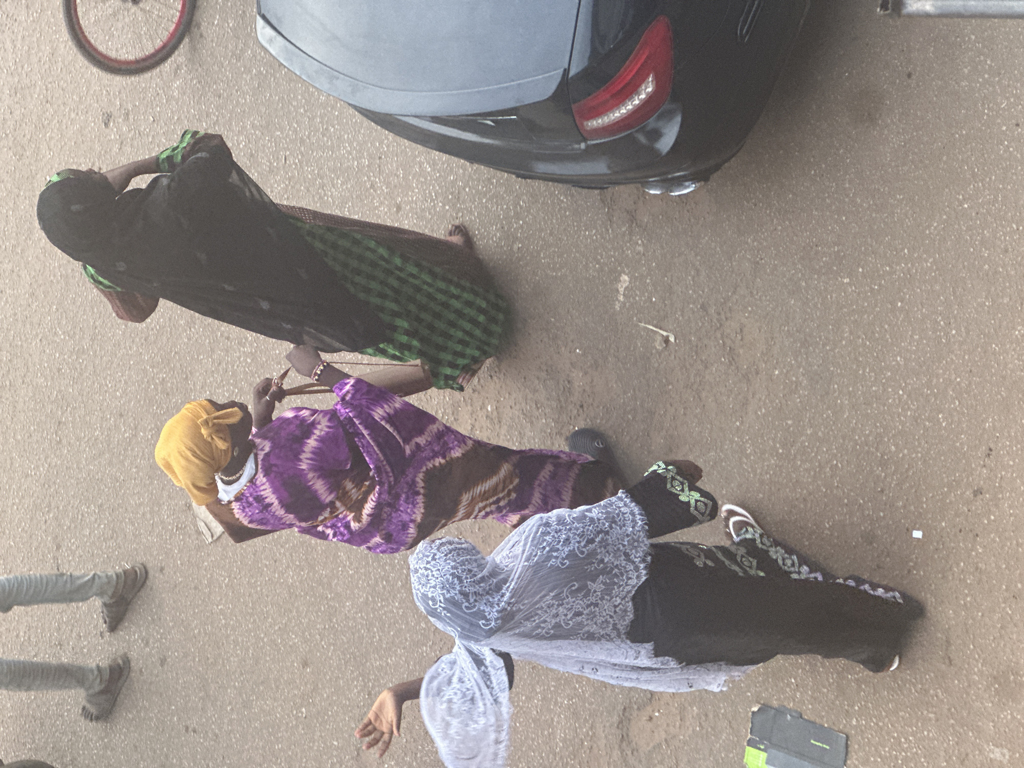 In Bukuru, Nigeria, three women are seen walking on a dirt road adjacent to a black car. The woman in the foreground is wearing a black garment with white and green embroidery and a white veil. To her right, a woman in a purple tie-dye dress and a yellow headscarf is walking. Behind her, a third woman is draped in a black hijab with green accents. The woman in the yellow headscarf appears to be holding a bag or purse. A bicycle wheel with a red rim is visible in the background, as is a partial view of the black car's rear. The ground is a mix of dirt and small stones, with some scattered debris.