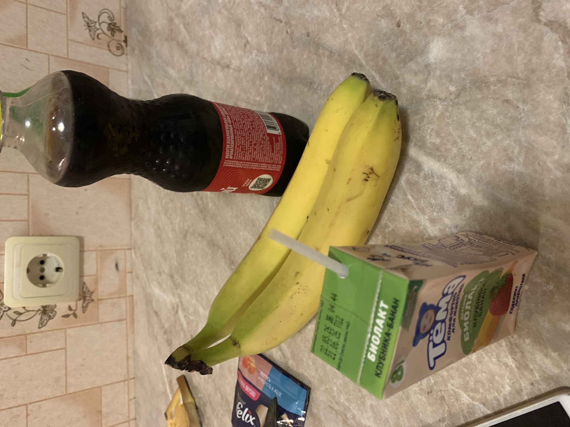 The image captures a collection of groceries and household items laid out on a patterned countertop, likely in a kitchen or dining area in Stavropol, Russia. The scene is brightly lit by an artificial light source.
The main subjects include a partially consumed plastic bottle of a dark, carbonated beverage, two ripe yellow bananas, and a small, rectangular juice box with a straw. In the lower left, a blue and silver pouch of pet food is partially visible, along with the corner of a smartphone. A white electrical outlet is mounted on a tiled wall in the background, which features a faint decorative butterfly and floral pattern.
Visible text on the beverage bottle's red label includes "НАПИТОК БЕЗАЛКОГОЛЬНЫЙ" (Non-alcoholic beverage) and "АРОМАТИЗИРОВАННЫЙ" (Flavored). The juice box is branded "Тёма" and identified as "ДЕТСКИЙ БИОЛАКТ" (Children's Biolact), specifically "КЛУБНИКА-БАНАН" (Strawberry-Banana) flavored, and notes that it "СОДЕРЖИТ ПРОБИОТИКИ" (Contains probiotics). Dates "22.05.26" and "21.06.26" are printed on its side. The pet food pouch is from the "Felix" brand and mentions "Лосось И ФОРЕЛЬ В ЖЕЛЕ" (Salmon and Trout in Jelly).