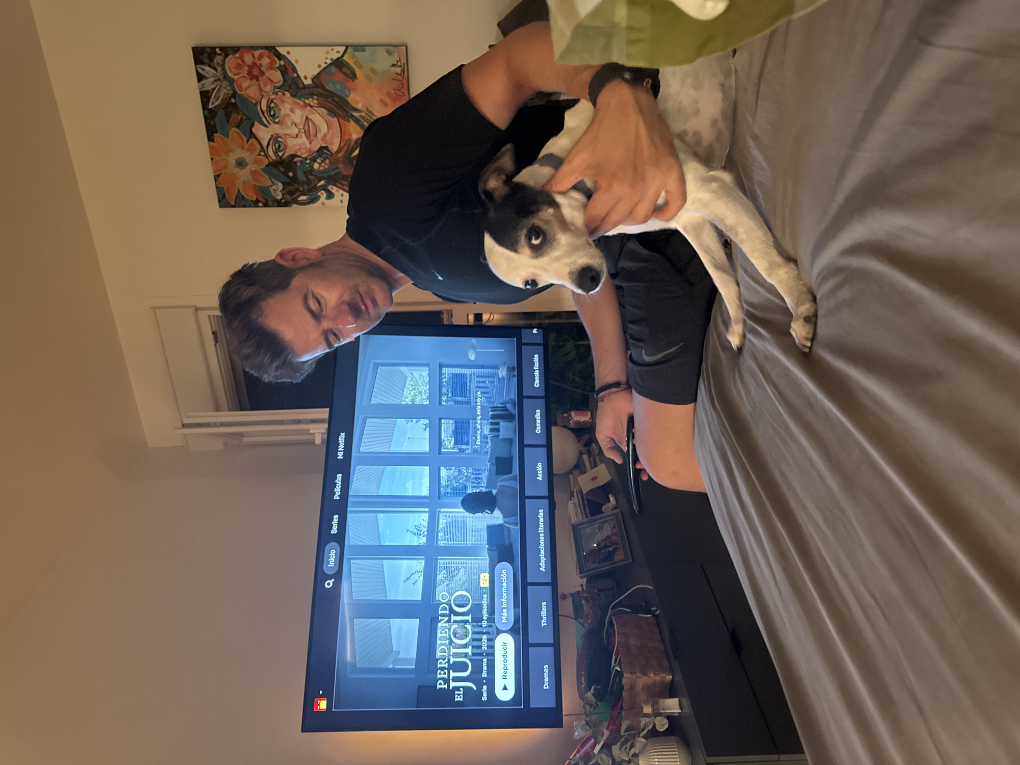 A man is holding a small, black and white dog on his lap while sitting in front of a television screen. The television is displaying a menu for a streaming service with options for "Inicio", "Series", "Películas", and "Mi Netflix". The man is wearing a black t-shirt and dark shorts, and the dog has a gray collar. The room appears to be a living room, with a bed in the foreground covered with a gray sheet. A painting of a woman with flowers in her hair is hanging on the wall in the background. The scene is illuminated by the glow of the television and ambient lighting. The overall impression is one of a relaxed evening at home.