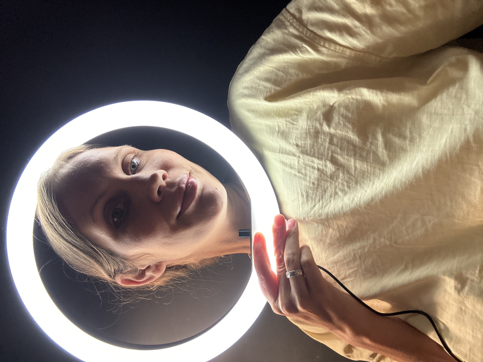 A woman is positioned in front of a ring light. She is fair-skinned, with blonde hair pulled back. Her left hand is visible holding the ring light's power switch, and she is wearing a silver ring. The background appears to be dark. Part of her light yellow shirt is also visible. It is unlikely that this image could be geolocated to Fethiye, Turkey without further information or context. The scene does not contain any identifiable landmarks or features associated with that specific location.
