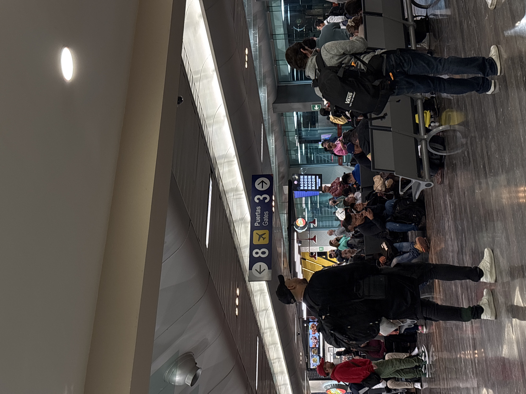 The image depicts a bustling indoor waiting area, strongly resembling an airport terminal. Many people are present, primarily adults, scattered across the scene. Most are seated on metal benches designed for waiting, while some individuals stand. In the immediate foreground, a person wearing a black jacket, black pants, and a black baseball cap stands facing away from the camera, with their back partially visible. Further back, another person in a grey hooded sweatshirt, jeans, and white sneakers stands by a row of seats, carrying a black backpack adorned with a "VANS" logo.
The setting features a modern architectural style with high ceilings and a polished, reflective dark floor that mirrors the overhead lighting and figures. Prominent overhead lighting includes strip lights running along ceiling structures and recessed circular lights. A blue vertical sign is visible in the mid-ground, indicating "Puertas 3 Gates" and displaying an airplane icon. Below this, the number "8" with a downward arrow suggests direction to a specific gate. An electronic display screen in the background repeatedly shows the word "BACON," likely an advertisement from a food vendor. The overall atmosphere suggests a typical travel waiting experience, with individuals patiently awaiting their departures.