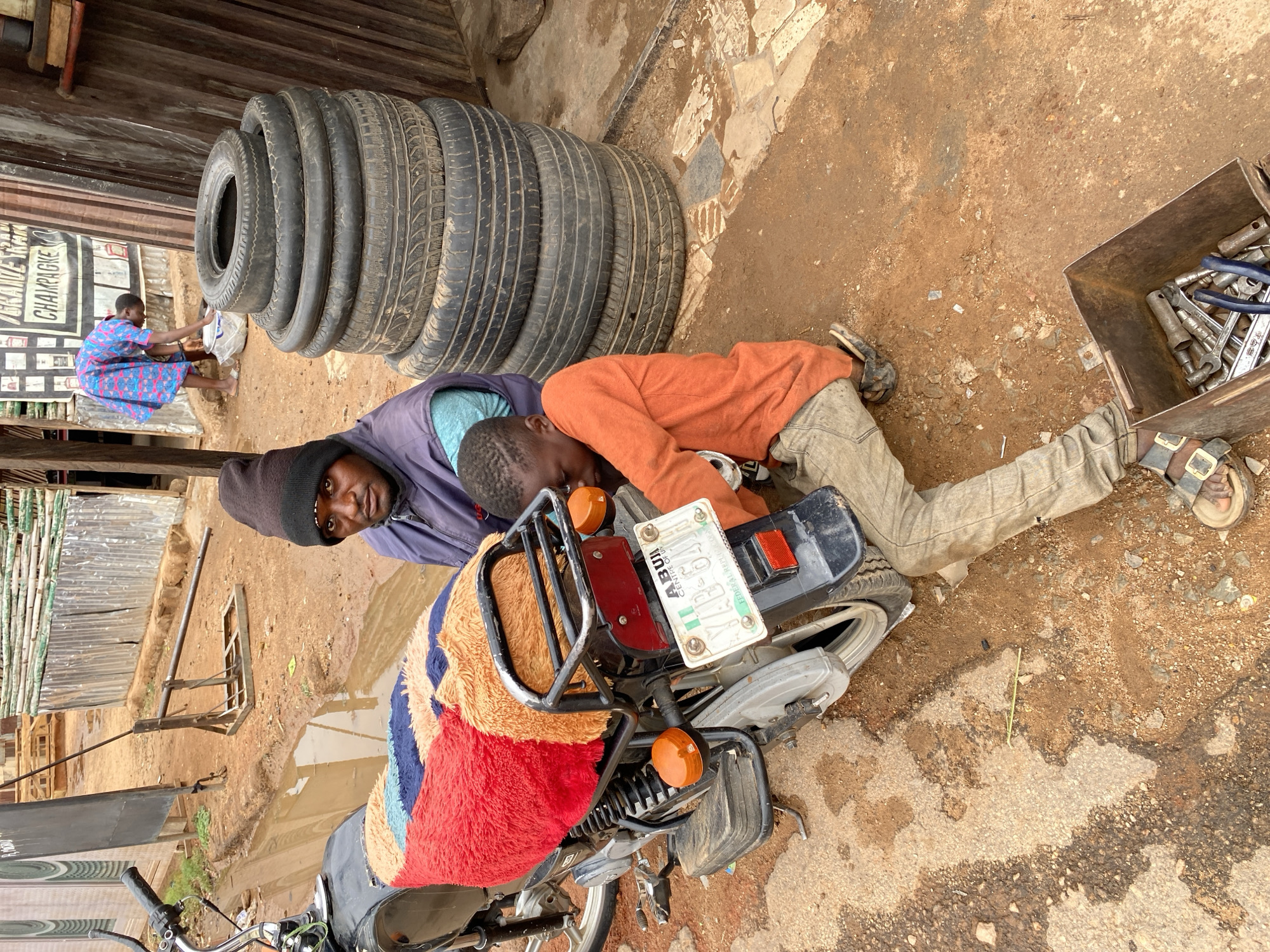 In Narayi, Nigeria, a man in a black beanie and blue jacket observes the viewer as a young boy in an orange shirt lies on his stomach, partially beneath a parked motorcycle. The motorcycle has a colorful cloth draped over the rear and a license plate reading "ABUI 04 AP". Next to the boy lies an open toolbox containing wrenches and other tools. A stack of used tires sits nearby. A woman in a blue dress is seated in the background near a building with writing on it. The ground appears to be dirt and concrete with puddles of water.