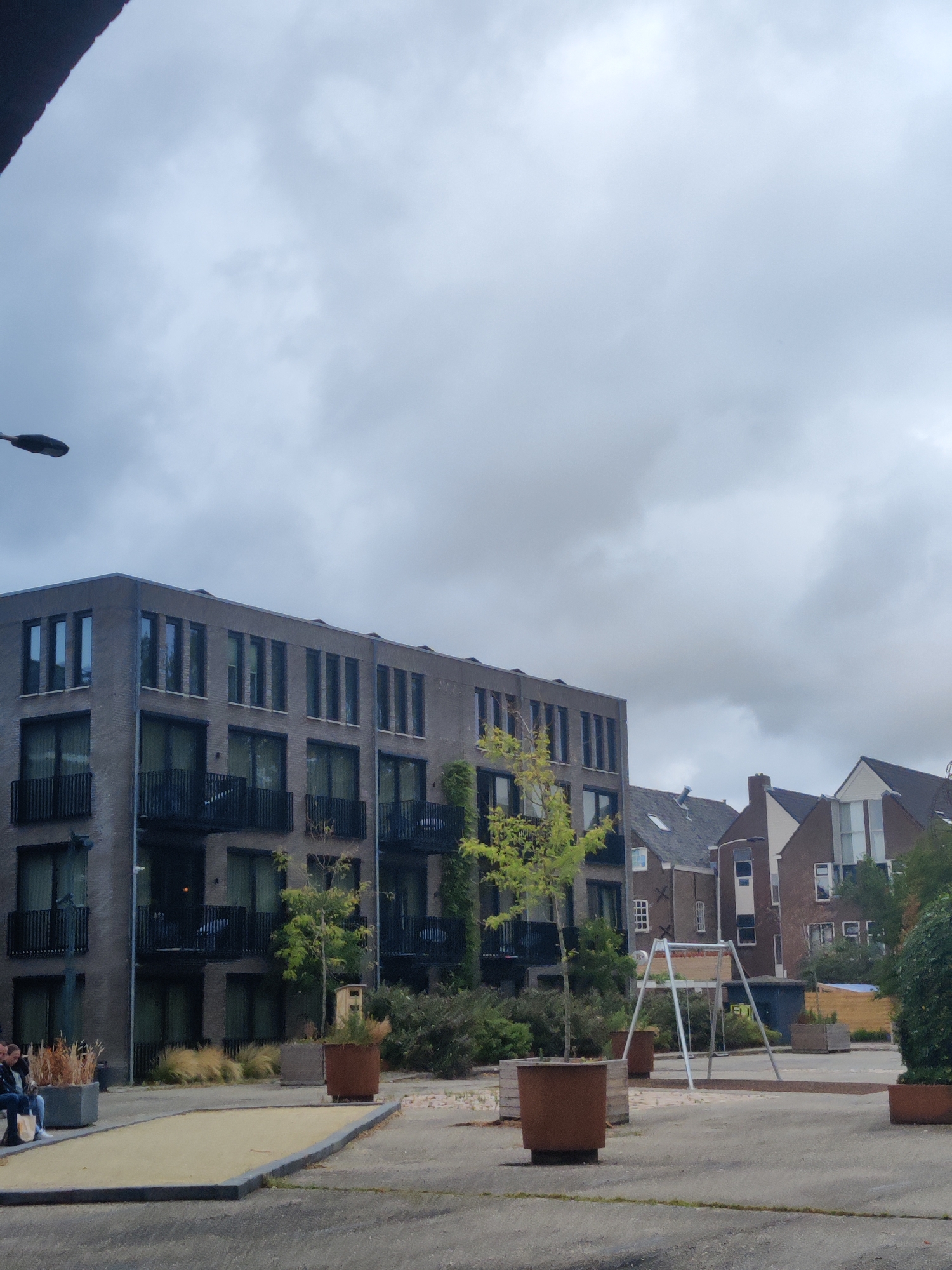 The image depicts an outdoor scene in the city of Delft, Netherlands, on a cloudy day. In the foreground, a paved area includes a bocce court with a yellow playing surface. To the left of the court, two individuals are seated on a bench, one wearing a dark jacket and the other a lighter colored top. Several large planters containing plants are scattered around the paved area.
Dominating the background is a modern, multi-story apartment building constructed with grey brick. It features numerous dark-framed windows and balconies. To the right of this building, a row of older, brick houses with pitched roofs are visible. A silver swing set is situated in the middle ground. The sky is overcast with heavy clouds.