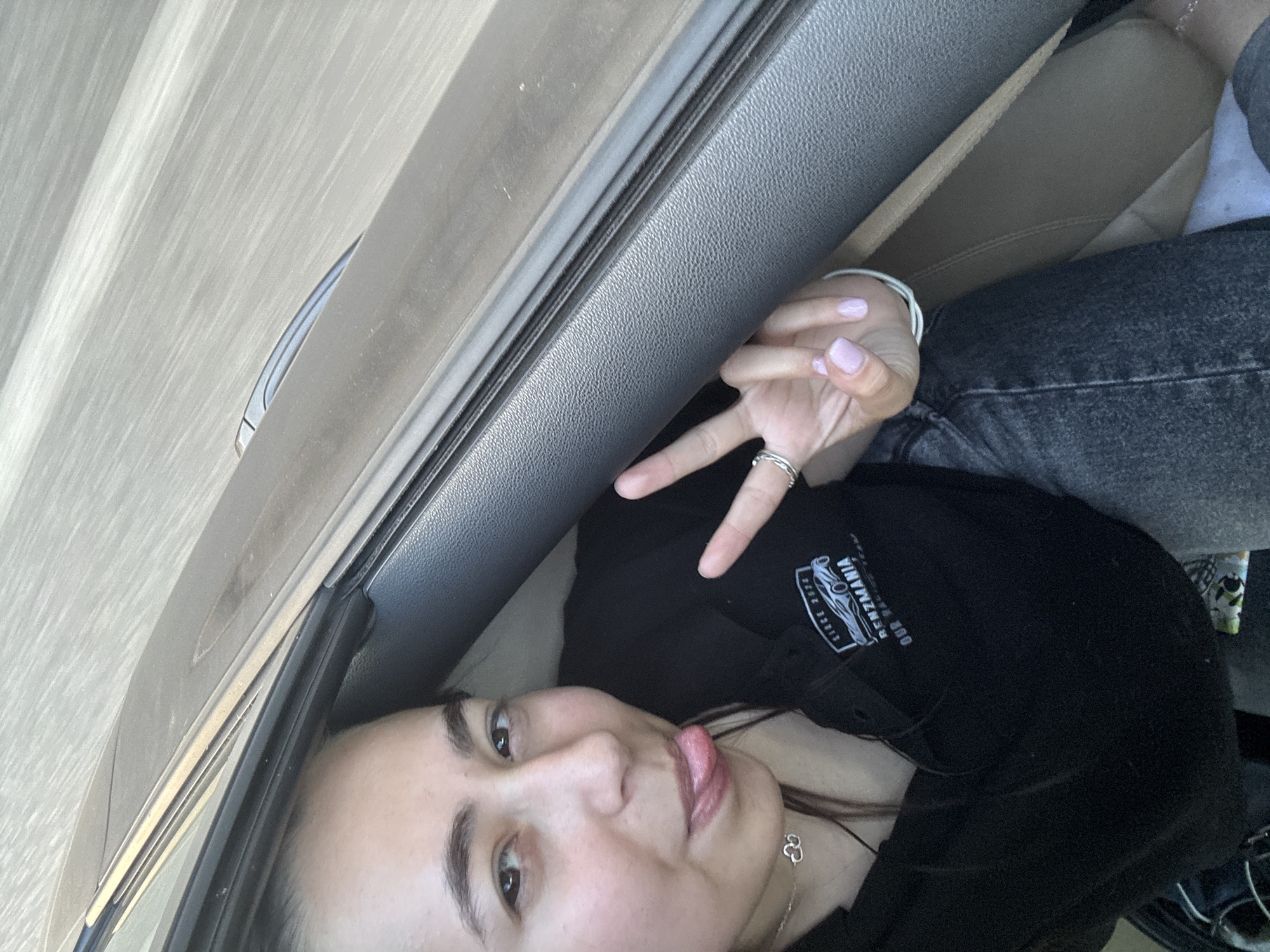 The image depicts a single individual, a young female, seated within the interior of a vehicle in Omarchevo, Bulgaria. She has dark hair, dark eyebrows, and light-colored skin. Her mouth is slightly open, and her tongue is extended in a playful gesture. Her left hand (viewer's right) is raised, forming a peace sign with her index and middle fingers. Her fingernails are painted a light lavender or pale pink. A ring is visible on her left ring finger, and a thin bracelet is on her left wrist. She wears a silver-toned necklace with a double cloud or interlocking heart pendant. The individual is dressed in a black collared shirt featuring a white logo that reads "SINCE 2024 BENZMANIA OUR RACING TEAM" alongside a stylized race car graphic. She is also wearing dark wash jeans. The car's interior shows dark grey textured plastic, likely a door panel or dashboard, and lighter tan or brown upholstery. The exterior view through the car window is blurred with horizontal streaks, suggesting the vehicle is in motion on a road.
