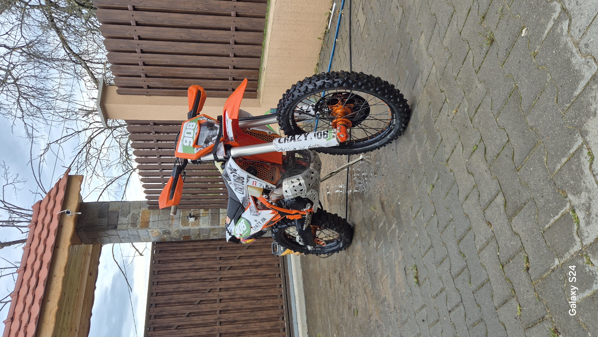The image displays an orange and white dirt bike, likely an enduro or motocross model, parked outdoors on a paved surface. The bike is positioned facing slightly right, with its front wheel angled towards the viewer. It features knobby tires, orange wheel hubs, and bright orange handguards and fenders. White body panels are adorned with black and green graphics, prominently displaying the text "CRAZY 106" and the number "906" (or "106" from different angles) on various parts, including the front number plate and side panels. A metal skid plate or engine guard is visible beneath the engine.
The setting appears to be a residential or private outdoor area, possibly a driveway or yard. The ground is paved with gray interlocking pavers, which appear damp in places, suggesting recent rain or washing. In the background, a dark brown wooden fence runs horizontally, behind which there's a light-colored building wall. To the left, a stone pillar supports a wooden roof structure with terracotta-colored tiles, forming part of a gate or entrance. Bare tree branches are visible against an overcast sky, suggesting cooler weather. A small white security camera is mounted on the stone pillar, and a blue hose is visible on the paved ground near the bike's front wheel. A white power outlet is on the wall behind the bike. The bottom right corner of the original image includes a "Galaxy S24" watermark.