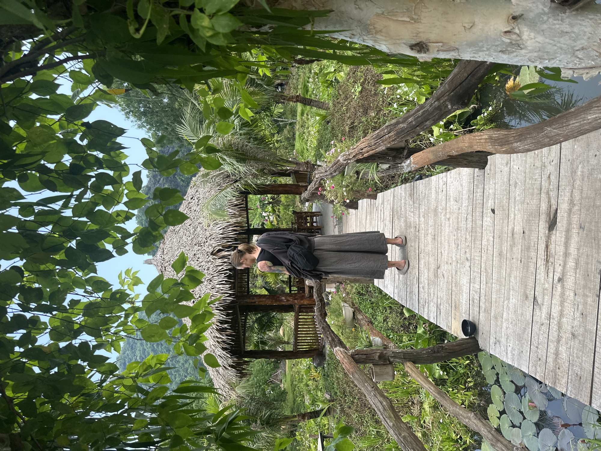 A woman with tattoos stands on a wooden bridge in a tropical garden setting, with a thatched-roof pavilion in the background. The woman is wearing a dark tank top and a long, flowing gray skirt, with a dark cloth draped over her shoulders and a black backpack. She is looking away from the camera, towards the pavilion. The bridge is made of weathered wooden planks and has rustic wooden railings. To the left of the bridge, there is a small pond with lily pads. The garden is lush and green, with palm trees and various plants. In the distance, there are mountains. The overall impression is one of tranquility and natural beauty. The location is in Nam Hoa Lư Ward, Vietnam.
