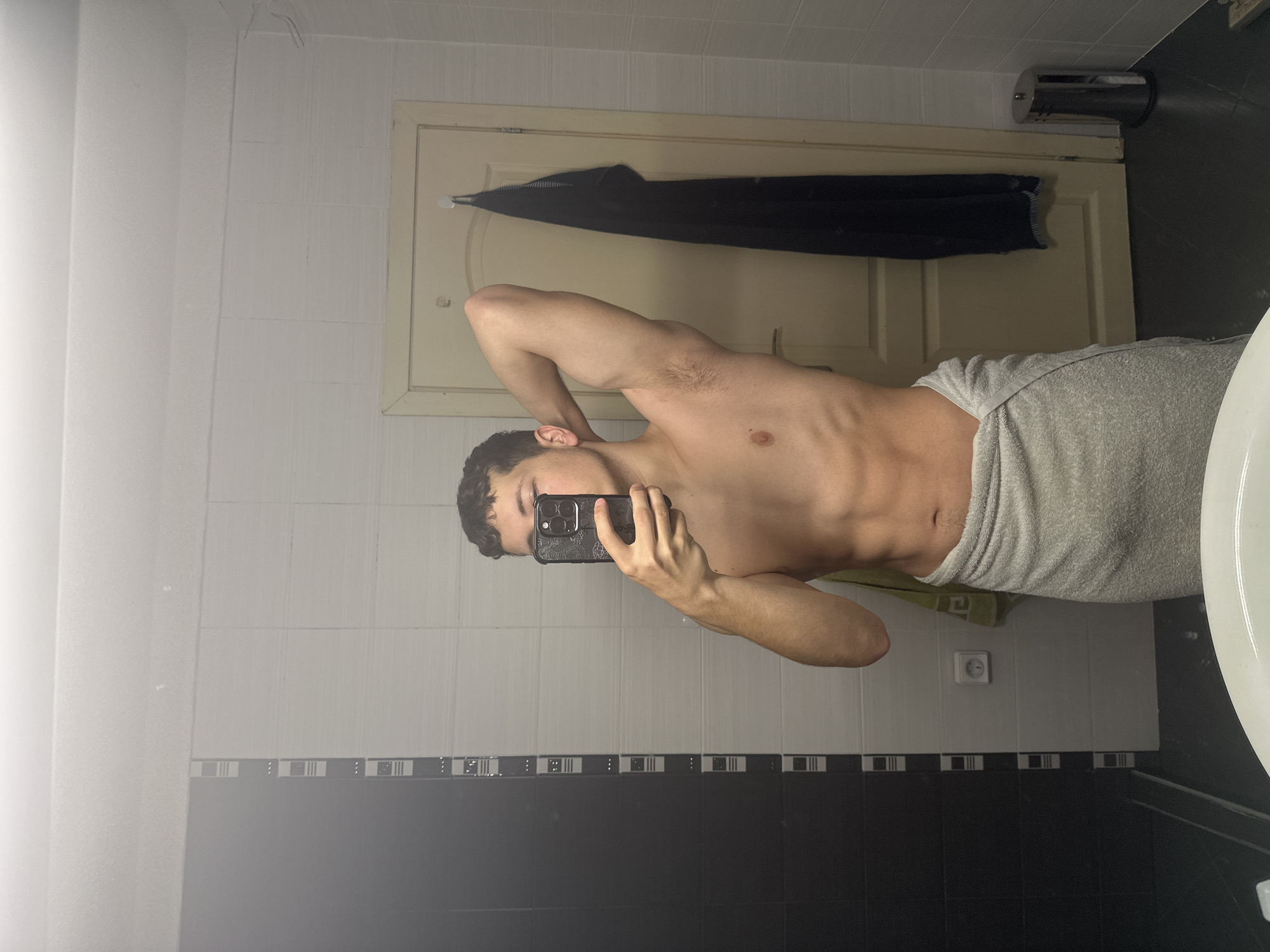 An indoor scene depicts a mirror selfie taken in a bathroom in the city of Unknown, Unknown. A youthful male individual stands shirtless, wrapped in a light grey towel around his waist. His right arm (viewer's left) is raised, holding a dark smartphone with a patterned case that obscures his nose and mouth. His left arm (viewer's right) is bent at the elbow, with the hand resting on the back of his head, revealing short, dark, wavy hair and visible armpit hair. His torso displays defined abdominal muscles.
The bathroom features a wall composed of large black rectangular tiles on the left, transitioning to white horizontal-textured tiles towards the center and right. A thin vertical strip of white tiles with small black rectangular insets separates the black and white sections. An electrical outlet is positioned on the white tiled wall to the lower left of the subject. A light green hand towel hangs on a hook to the right of the outlet. Centrally in the background is a light-colored (beige/cream) wooden door, featuring a white adhesive hook from which a dark blue or black striped towel hangs vertically. To the far right, a cylindrical stainless steel trash can with a pedal mechanism rests on the dark tiled floor. The bottom edge of a white porcelain sink basin is visible in the foreground. The ceiling is white and undecorated.