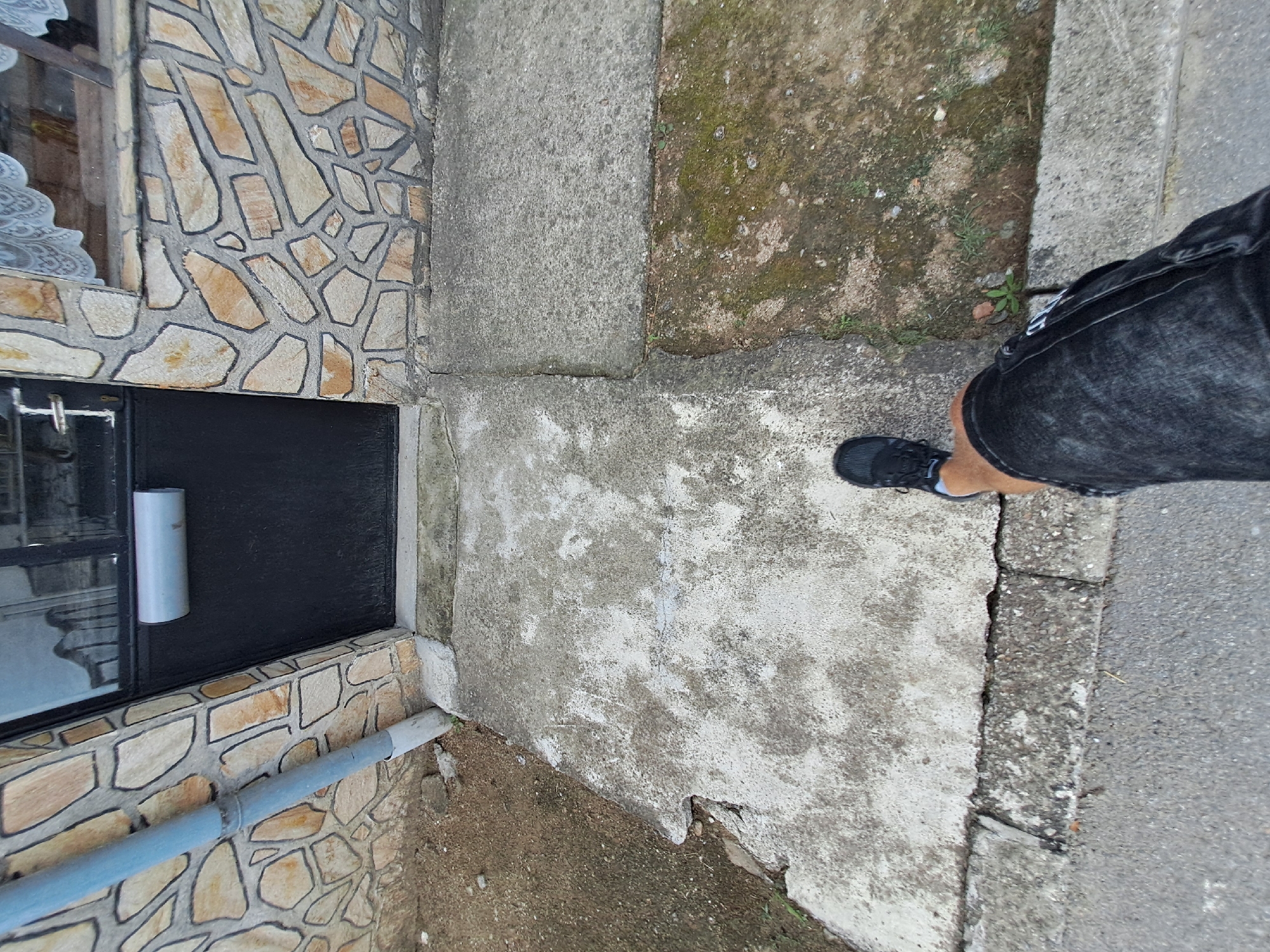 A person's legs and feet are visible in the lower right quadrant of the image, wearing black denim shorts and black sneakers. The individual is standing on a concrete surface that transitions to a patch of dirt and moss. To the left of the concrete is a section of wall adorned with irregular, light-colored stones outlined in dark grout, forming a mosaic-like pattern. Below this stone wall, a black rectangular area is visible, possibly a window or a door. A grey pipe runs horizontally along the bottom of the stone wall. The scene appears to be outdoors, with natural light suggesting daytime. The overall setting is likely an exterior walkway or path next to a building.