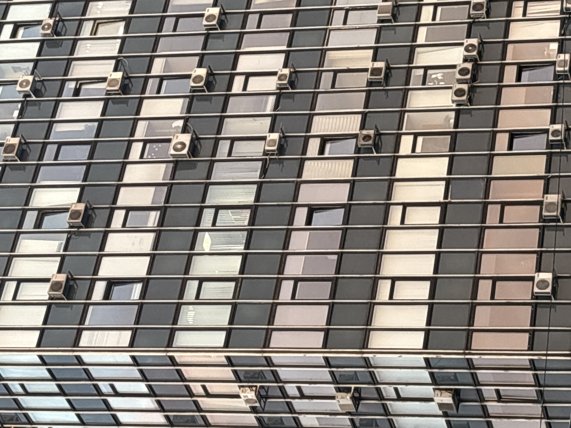 This is a medium shot of the exterior of a modern, multi-story building in an urban setting. The building is constructed with a grid of dark gray panels and numerous windows. Many of the windows are rectangular and appear to be made of glass, reflecting the sky and surrounding environment. Several rectangular, white outdoor air conditioning units are mounted on brackets outside the windows on various floors. The scene is illuminated by natural daylight, suggesting it was taken during the day. There are no people visible in the image.