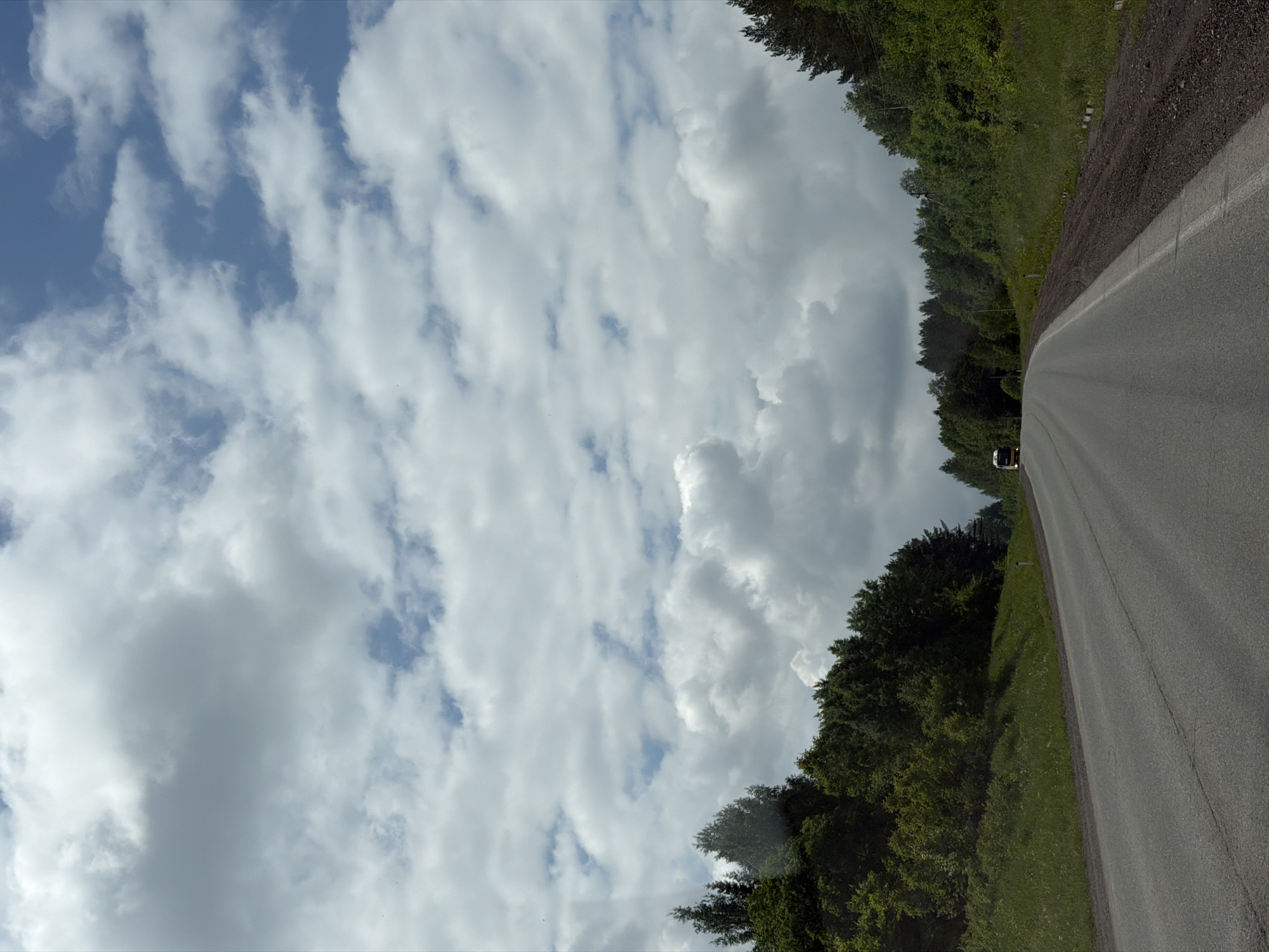 The image displays a rural road scene, captured from a slightly elevated perspective, with the original image appearing rotated 90 degrees clockwise. When viewed upright, the scene features a two-lane asphalt road curving gently into the distance on the right side of the frame, bordered by a dirt and gravel shoulder. A single white line is visible on the asphalt, likely marking the edge of the lane or shoulder.
To the left of the road, dense green trees and foliage form a thick forest that stretches towards the horizon. The trees appear lush, indicating a temperate climate. A small, light-colored vehicle, possibly a white van or truck, is visible on the road in the mid-ground, heading away from the viewer.
Above the tree line and road, the sky is filled with a mix of cumulus and stratocumulus clouds. Patches of clear blue sky are visible between the clouds, suggesting a partly cloudy day rather than an overcast one. The lighting indicates it is daytime. No specific time of day can be determined beyond daylight hours. There is no visible text or identifiable people in the image. The overall impression is one of a peaceful, natural landscape along a country road in Башкултаево, Russia.