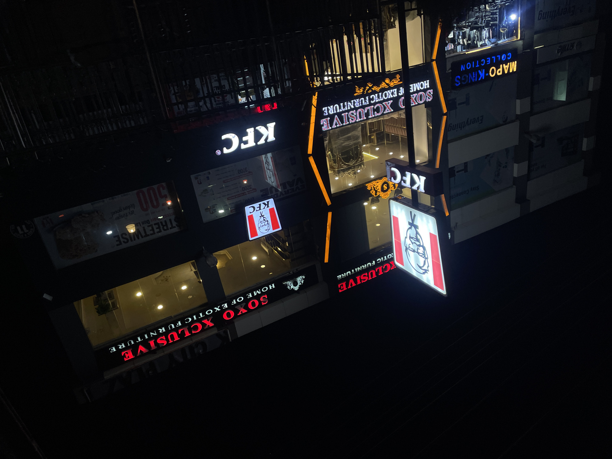 This image captures a bustling commercial street in Abuja, Nigeria, at night, viewed from a tilted perspective looking upwards at several multi-story buildings. The scene is dominated by brightly illuminated signs against the dark sky, indicating active businesses.
The most prominent establishment is a KFC restaurant, identified by its iconic red and white logo featuring Colonel Sanders and the distinct "KFC" lettering, which appears on multiple signs. One sign clearly displays the brand's slogan, "it's finger lickin good." Adjacent to and above the KFC, a furniture store named "SOXO XCLUSIVE HOME OF EXOTIC FURNITURE" is prominently featured with large, glowing red and orange signs. Through its expansive windows, intricate furniture pieces, including ornate chairs and decorative items, are visible, lit by interior lights.
Further along the street, another business, "MAPO-KINGS COLLECTION," is identifiable by its blue and yellow illuminated sign. The word "Everything" appears on several signs on the right side of the image, potentially indicating a general merchandise store or another retail outlet. A sign for "STREETWISE" is also visible, along with numerical values like "500," "2760," "3200," and "3700," which may represent prices or special offers. Other discernible texts include "VALVE," "smile," "Stay Connecte," and "K CITY PL." The overall impression is one of a vibrant, active urban commercial area operating after sunset.
