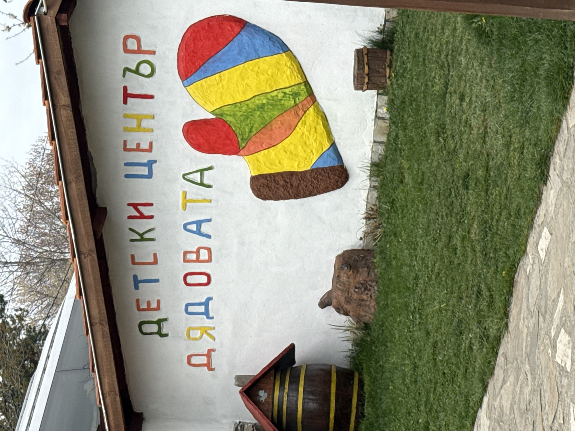 The image shows the facade of a building in Zlatitsa, Bulgaria. The facade is painted white with a large colorful mitten and the words "Детски център Дядовата" (Children's Center Grandfather's) written vertically in different colors. Next to the building is a small patch of grass and a stone walkway. There are also some decorative wooden objects.