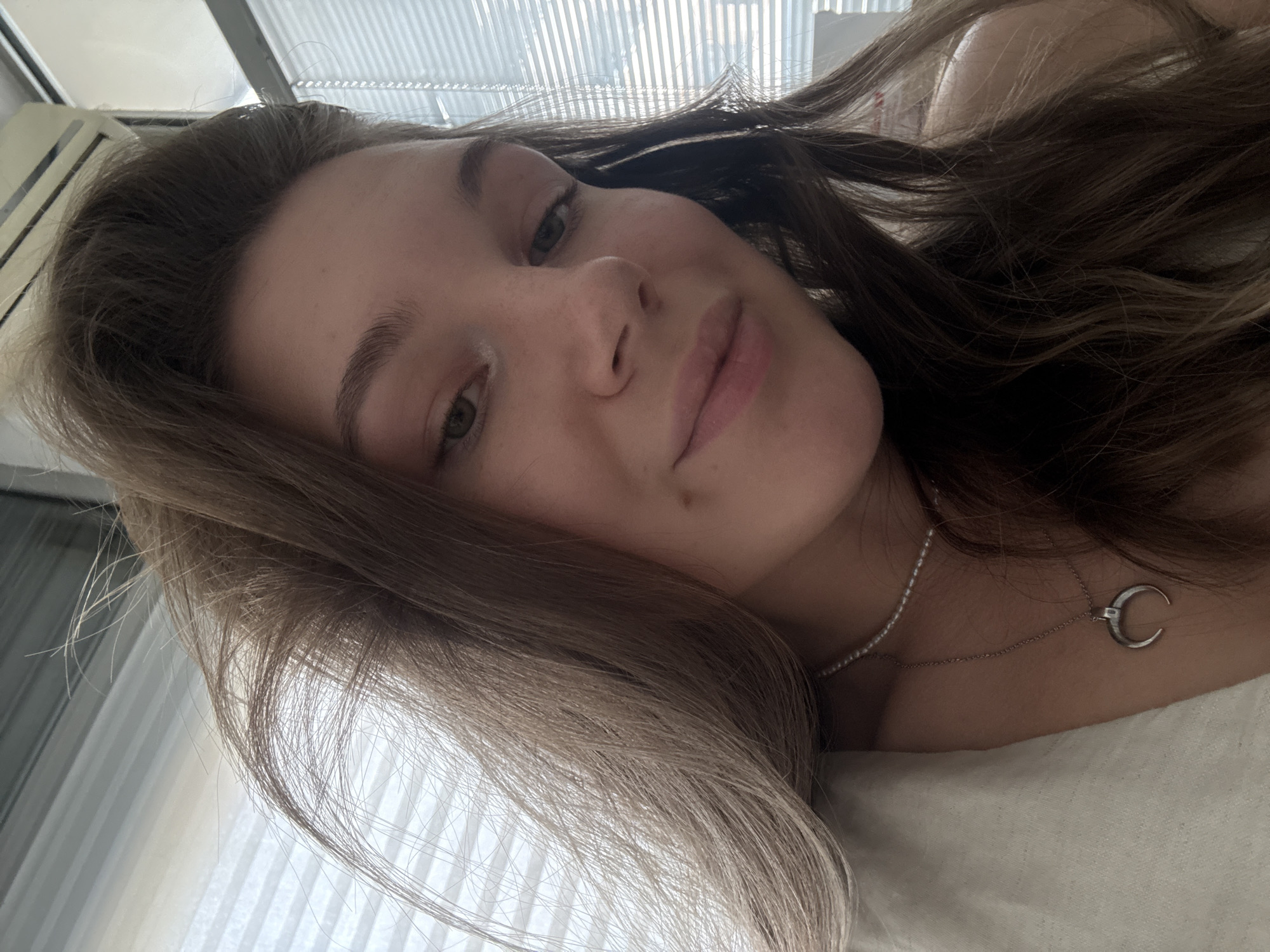 A close-up, selfie-style shot features a young woman with long, light brown hair, styled with volume, framing her face. She is wearing a light-colored garment and a delicate pearl necklace with a crescent moon pendant. Her facial features include green eyes, a subtle smile, and a dimple on her right cheek. The background suggests an indoor setting, possibly an apartment in Vracar Urban Municipality, Serbia, with a window covered by horizontal blinds and an air conditioning unit visible at the top. The lighting appears natural, coming from the window. The overall impression is casual and relaxed.