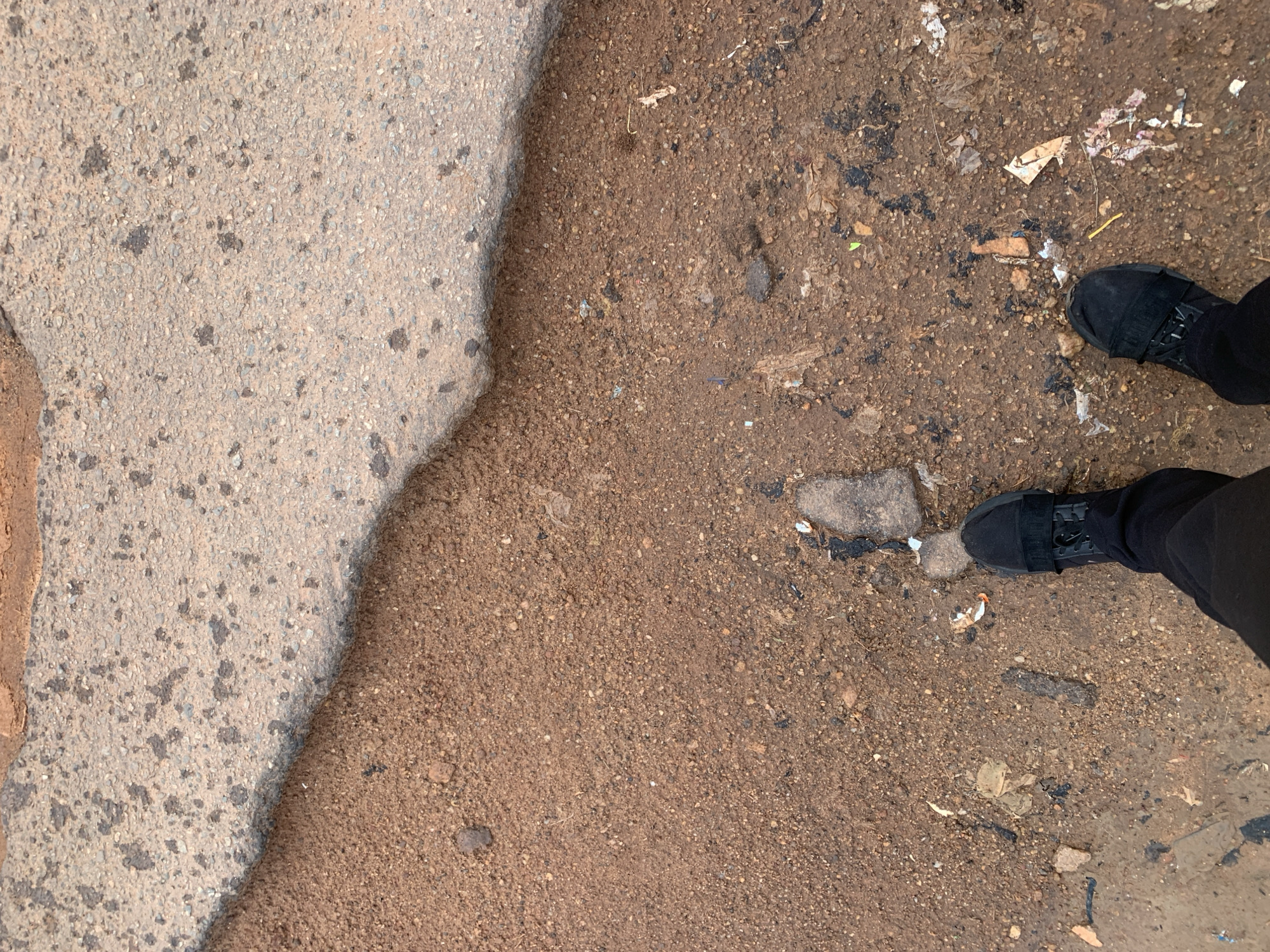 A first-person perspective shot looks down at a person's feet, clad in black sneakers and pants, standing on a patch of dirt and debris next to a worn asphalt surface. The asphalt, a light grey with dark speckles, has a rough, uneven texture and appears to be broken or eroded along the edge where it meets the dirt. The dirt itself is a reddish-brown, loose and granular, with scattered small stones, bits of trash, and dark stains. The ground is uneven and appears to be outdoors.
