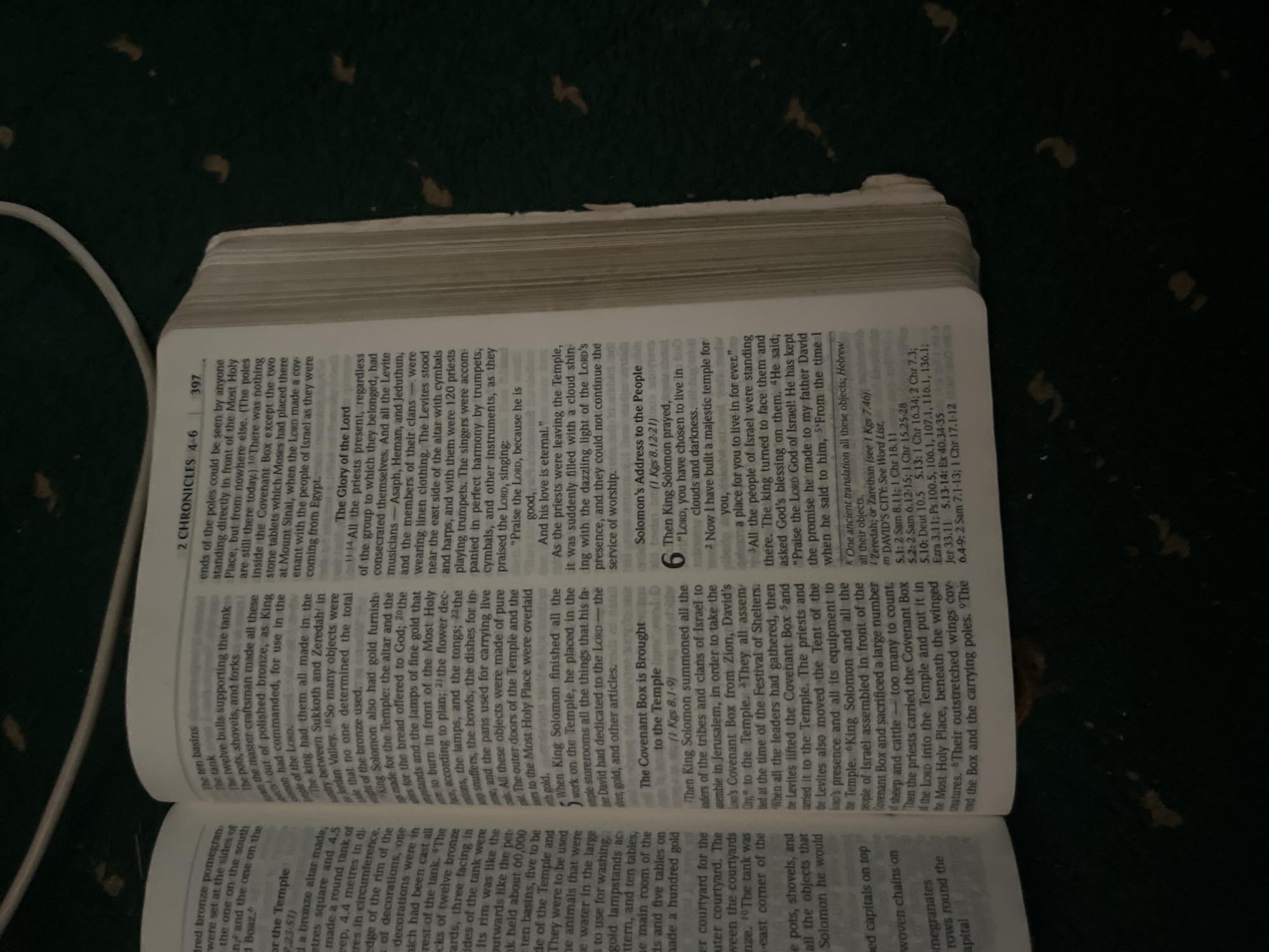 The image is an overhead close-up of an open Bible, showing text from 2 Chronicles 4-6 and part of chapter 7 on the right page, and text from the previous chapter on the left page. The Bible appears to be worn with age and use, with dog-eared pages and some yellowing. The lighting is dim, and the background is a dark, textured surface, possibly a carpet or fabric, with some blurry, scattered spots of light. The text is primarily in English, with some Hebrew script visible in a footnote. No people are visible in the image. The scene is indoors and appears to be a still-life composition focused on the book. There is no information in the image to suggest any connection to Jauro Yinu, Nigeria.