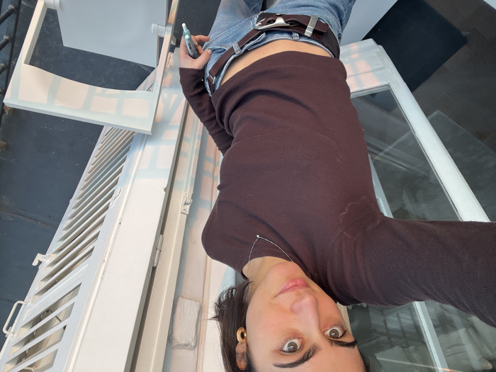 A young adult female subject, with dark hair and eyes, is depicted in a waist-up, self-taken portrait on a Parisian balcony. She wears a dark brown long-sleeved top with visible sweat stains under the armpits, light blue jeans, a dark brown belt with a metallic buckle, a thin silver necklace with a small pendant, and gold hoop earrings. She holds a silver and white cylindrical object, possibly a vape pen or marker, in her right hand near her hip. The subject looks directly at the camera with a slight smile. Behind her to the left is a white-framed glass French door or window reflecting light. To her right, a white louvered shutter, consistent with Parisian architecture, is partially open. A light-colored outdoor chair is visible in the lower right foreground. The scene is illuminated by natural light, suggesting either late afternoon or early morning.