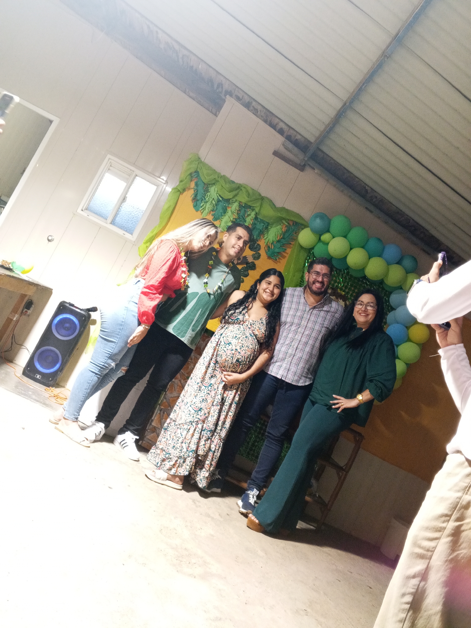 A group of six people are posing for a photo in an indoor location in the city of Unknown, Unknown. The group consists of four women and two men. Two of the women are visibly pregnant. One of the women is wearing a red shirt and jeans. The other woman is wearing a patterned dress. One of the men is wearing a green shirt and the other man is wearing a plaid shirt. The other woman is wearing a green shirt and pants. The sixth person taking the photo is wearing a white shirt and tan pants. Behind the group is a green and blue balloon arch and a backdrop with green decorations. There is a speaker on the left side of the image. The location is a room with white walls and a window.