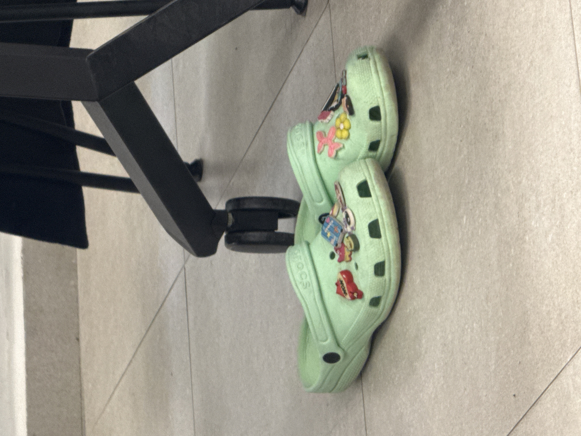 The scene presents a pair of light green Crocs shoes placed on a tiled floor, possibly inside a building in Tel-Aviv, Israel. The Crocs are decorated with Jibbitz charms. Part of a black office chair's base is visible near the shoes, suggesting an indoor setting. No people are visible in the image. The environment is clean and well-lit.