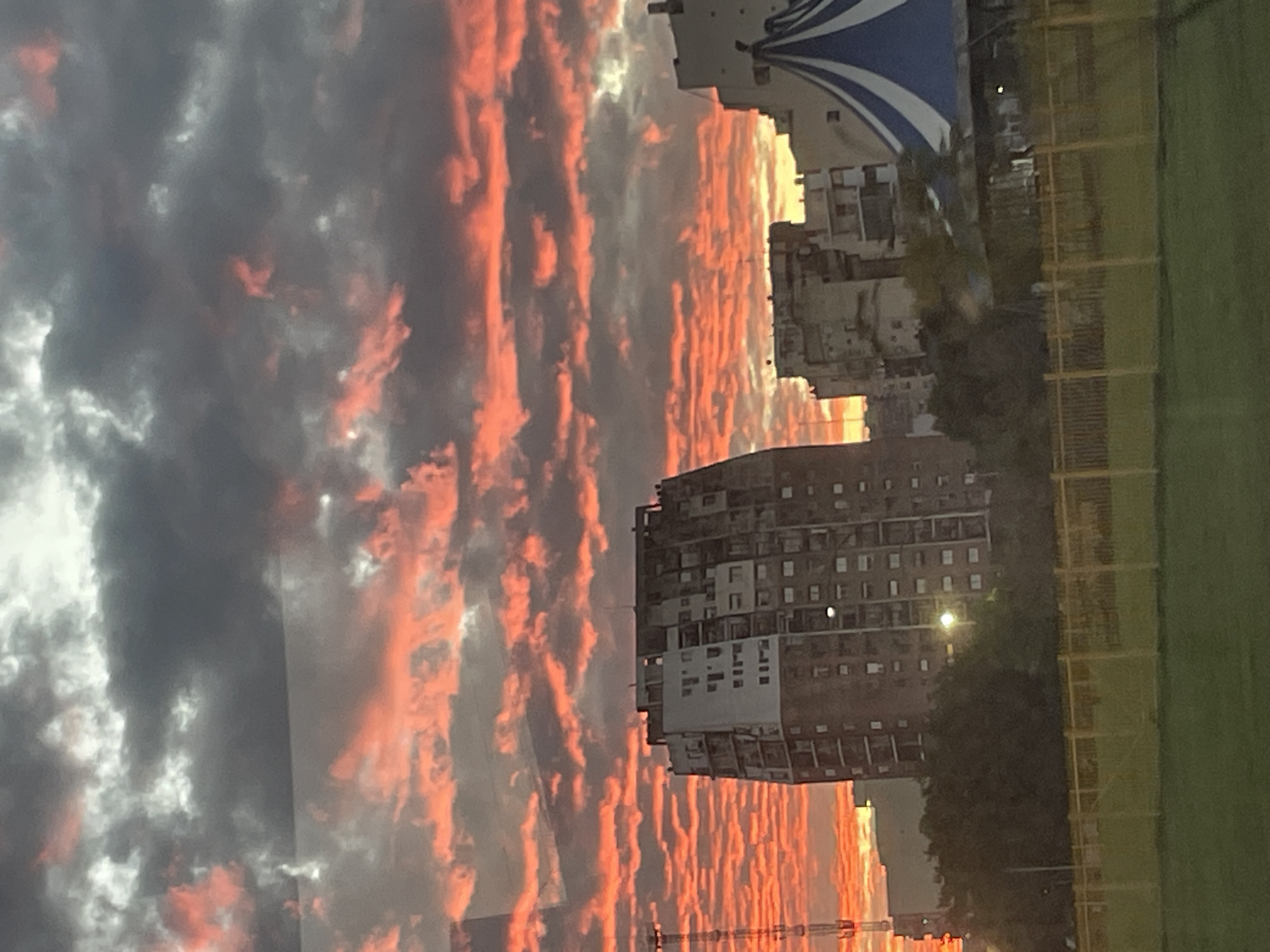 Here's a description of the image:
The image depicts a dramatic, colorful sunset in an urban environment, likely in Buenos Aires, Argentina. In the foreground, there is a green field with a yellow fence, suggesting a park or recreational area. Beyond the field, there is a tall, grey apartment building, possibly typical of Buenos Aires architecture. In the background, a mix of low-rise buildings and what appears to be a blue and white striped tent-like structure. The sky is the focal point, filled with dark, heavy clouds illuminated by the sunset, creating streaks of orange and red. There are power lines in the foreground. Overall, the scene captures the contrast between urban life and natural beauty.